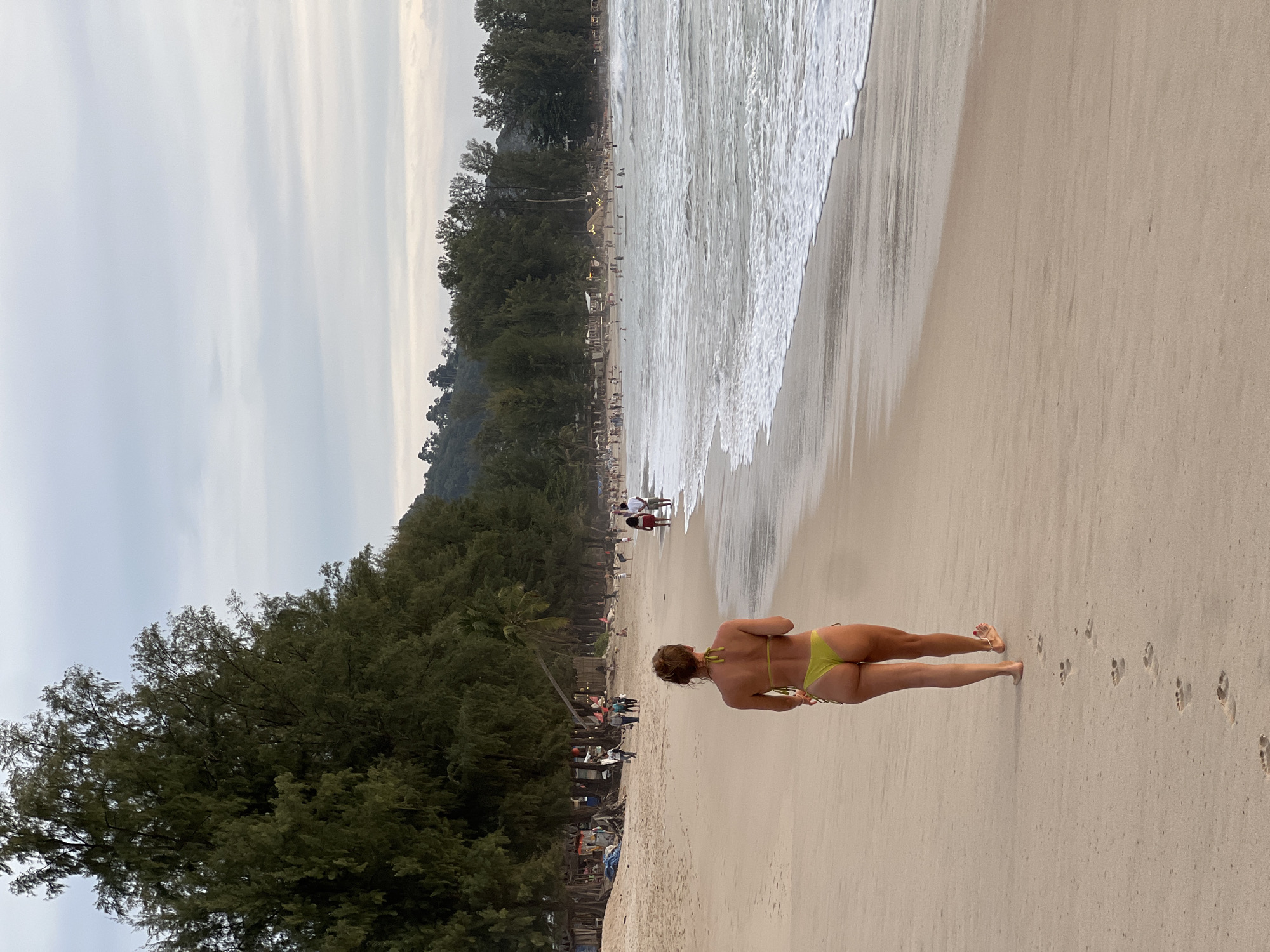 A woman in a lime green bikini walks away from the camera on a sandy beach in Choeng Thale, Thailand. Her footprints are visible in the sand behind her. The ocean with waves is to her right, and a line of trees and some buildings are visible further down the beach. Several other people are scattered along the beach and in the water. The sky is overcast.
