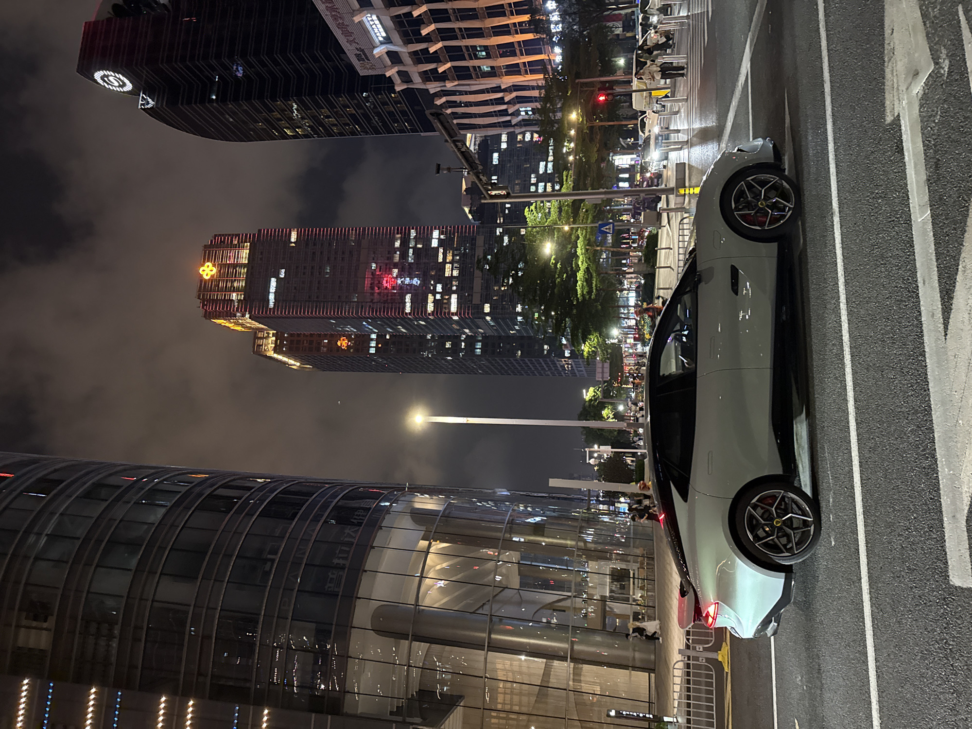 The image depicts a nighttime scene in Futian District, China. The foreground features a white sports car parked on an asphalt street with painted white lines. A modern building with a curved glass facade is visible to the left. In the background, illuminated skyscrapers rise against a cloudy sky. A street light casts a glow. Pedestrians can be seen on the sidewalks adjacent to the buildings. Trees line the street, with the streetlights illuminating them. The image focuses on the modern urban architecture of the district.