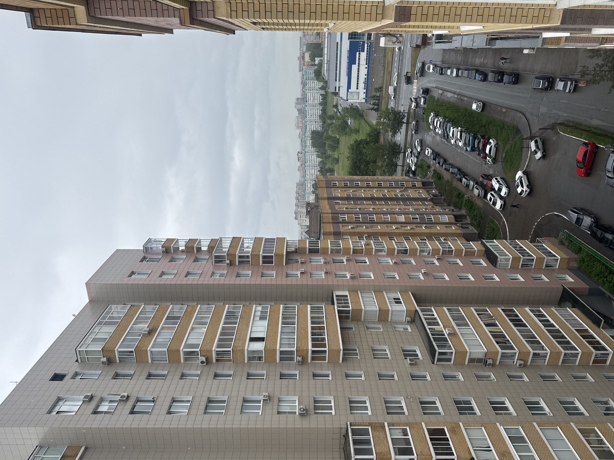 View of a city from an elevated position, likely a high-rise building. Overcast sky. Below are more buildings, a road with parked cars, and green spaces. The buildings in the foreground are multi-storied residential complexes with tiled exteriors and numerous windows, some with air conditioning units. The road has a line of parked cars along the curb. In the distance, other buildings and structures are visible.