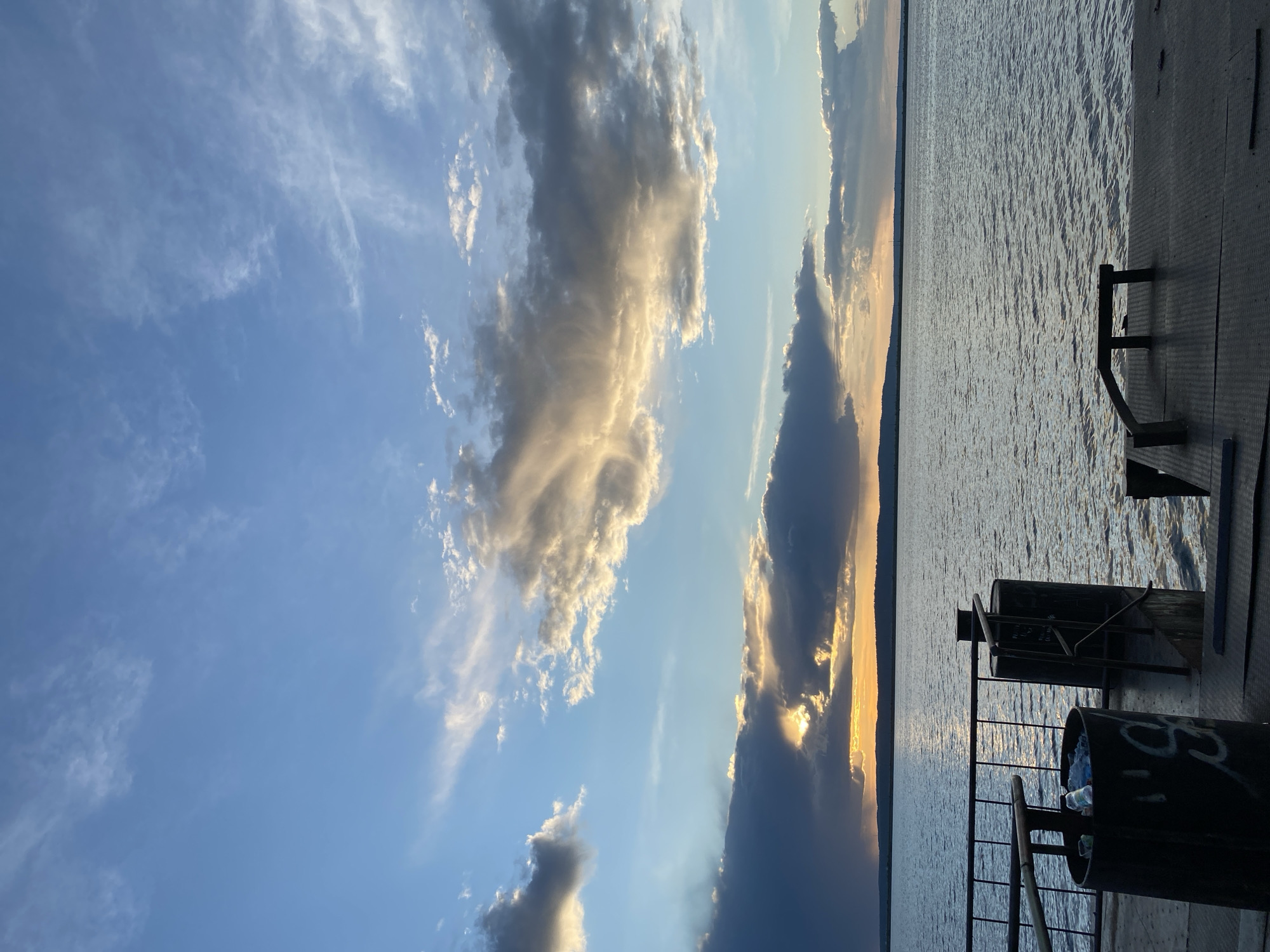 The image captures a sunset view from a dock or pier overlooking a body of water, likely the Kama River. The location is near Chaikovsky, Russia, indicated by the geography and architecture. The sky displays a gradient of blue to golden hues with scattered cumulus clouds illuminated by the setting sun. On the right side, the metallic deck and railing of the pier are visible. A garbage can sits near the railing. The water surface shows subtle ripples reflecting the colors of the sky. A treeline can be seen in the distance.