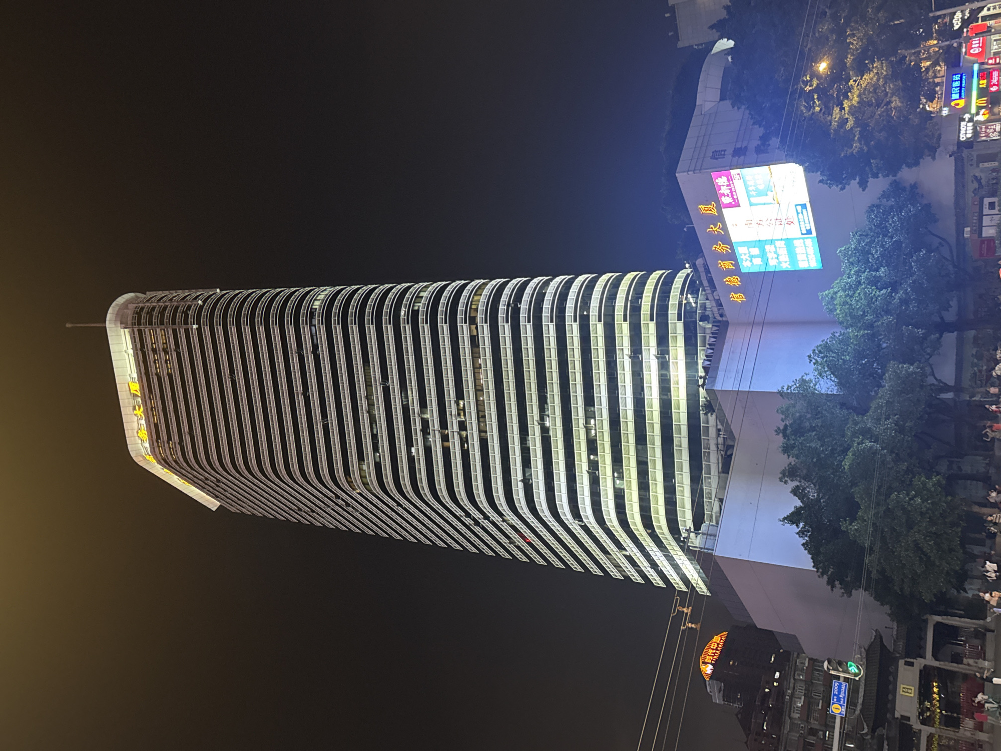 At night, a tall, modern building with a curved, striped façade dominates the view. It is illuminated, casting light onto the dark sky. To the right, smaller buildings and a brightly lit commercial complex are visible. Streetlights and illuminated signs add to the nighttime ambiance. Trees are interspersed among the buildings. The scene includes elements typical of an urban environment. Several signs, including "南方公证处", "信德商务大厦", and "健民医药" can be seen, indicating the location is in Yuexiu District, China. People can be seen indistinctly in the lower right corner of the image.