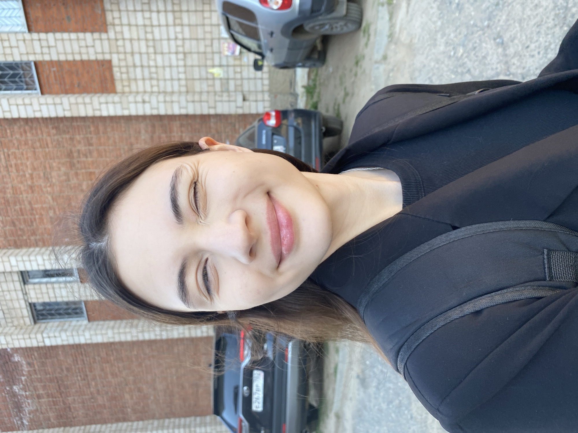 A woman is standing outside in what appears to be a city environment. She is wearing a black jacket over a black top and a black backpack. A brick building with white and brown brick patterns is visible in the background. Several cars, including a dark gray compact car, a dark-colored SUV, and a dark sedan with a visible license plate "E267BP 782," are also visible. The image seems to be taken in Votkinsk, Russia, given the license plate corresponds to the Russian Federation.