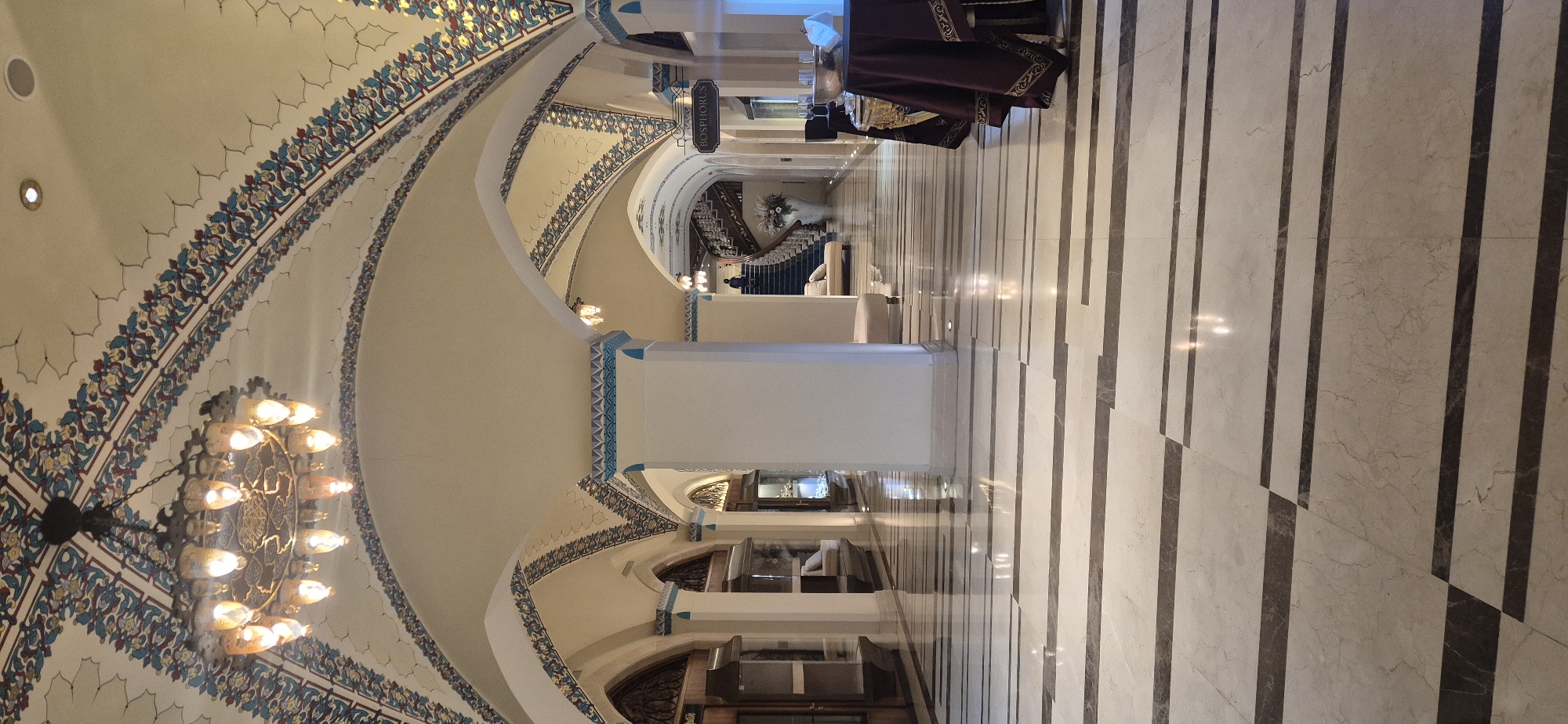 The image depicts the interior of a grand hallway with an arched ceiling adorned with intricate floral patterns in blue, yellow, and red. A large, ornate chandelier hangs from the center of the ceiling, illuminating the space. The walls are a light color, and white pillars support the arched structure. The floor is a polished marble with a pattern of alternating light and dark stripes.
To the left, glass display cases are visible, suggesting a museum or exhibition space. Further down the hallway, a grand staircase with a dark railing curves upwards, and a large white vase holding a floral arrangement is placed at its base. A person in silhouette can be seen on the stairs. A sign reading "Bosphorus" hangs from the ceiling on the right side of the hallway. In the foreground on the right, a table draped with a dark red cloth and adorned with gold trim is set up, with glasses and a bowl on top, possibly for an event. The lighting is warm and atmospheric.
This scene is likely located at the Aksu Museum in Aksu, Turkey, given the "Bosphorus" sign which may indicate a connection to Istanbul or a theme within the museum. The overall impression is one of opulence and historical significance.