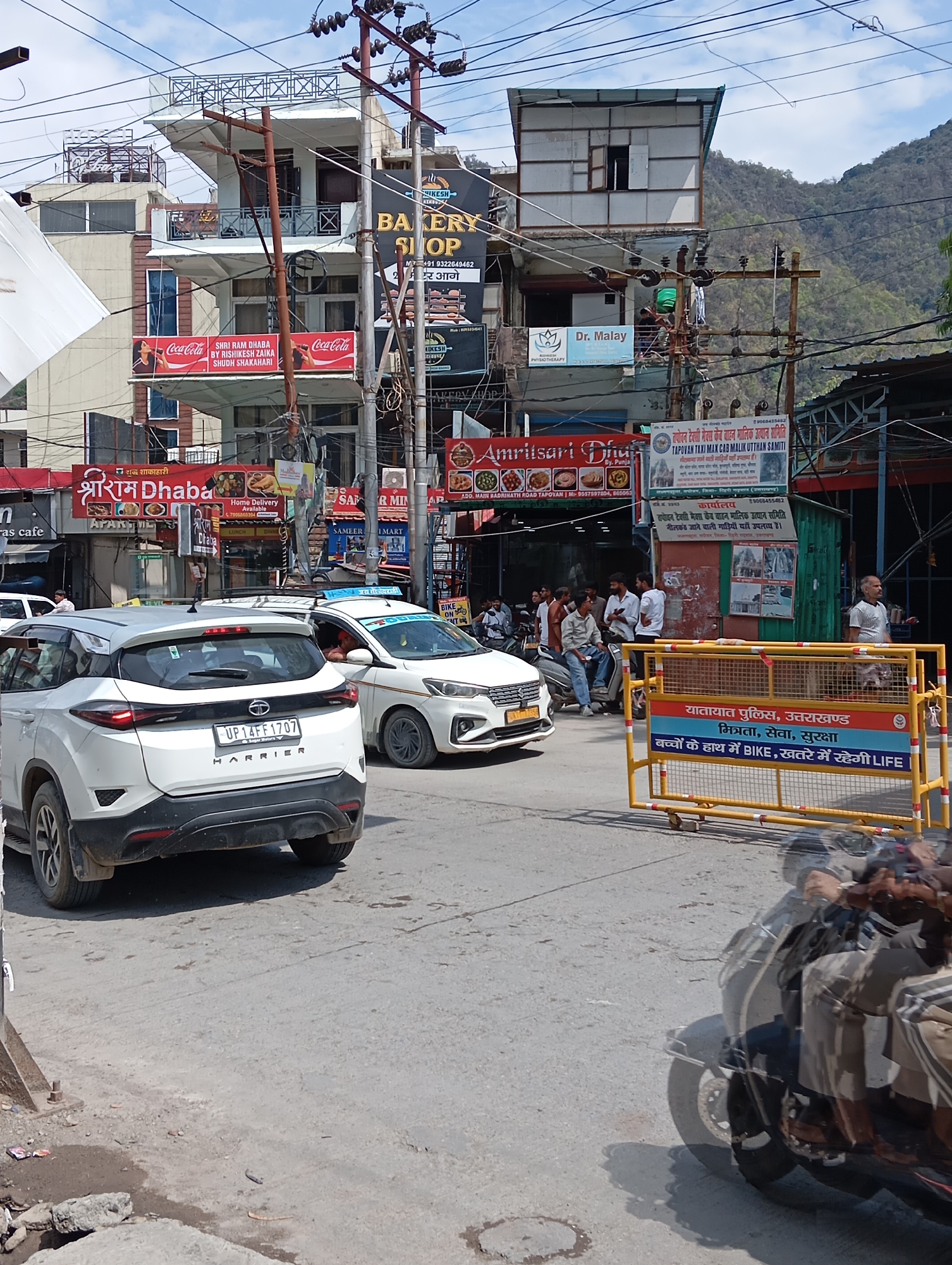The image captures a bustling street scene in what appears to be Jonk, India, based on the signage and vehicle registration. A white Tata Harrier SUV with license plate "UP14FF1707" is prominently displayed in the foreground, partially obscuring a white taxi. Several people are gathered near a row of businesses, including "Amritsari Dhaba" and "Shri Ram Dhaba." The buildings feature a mix of architectural styles and are adorned with numerous signs and advertisements, including Coca-Cola and a bakery shop. Overhead, a complex network of electrical wires crisscrosses the street. A yellow traffic barricade bearing a safety message in Hindi concerning children and bikes partially blocks the view of a group of people and a scooter. In the background, a lush green hillside rises behind the buildings.