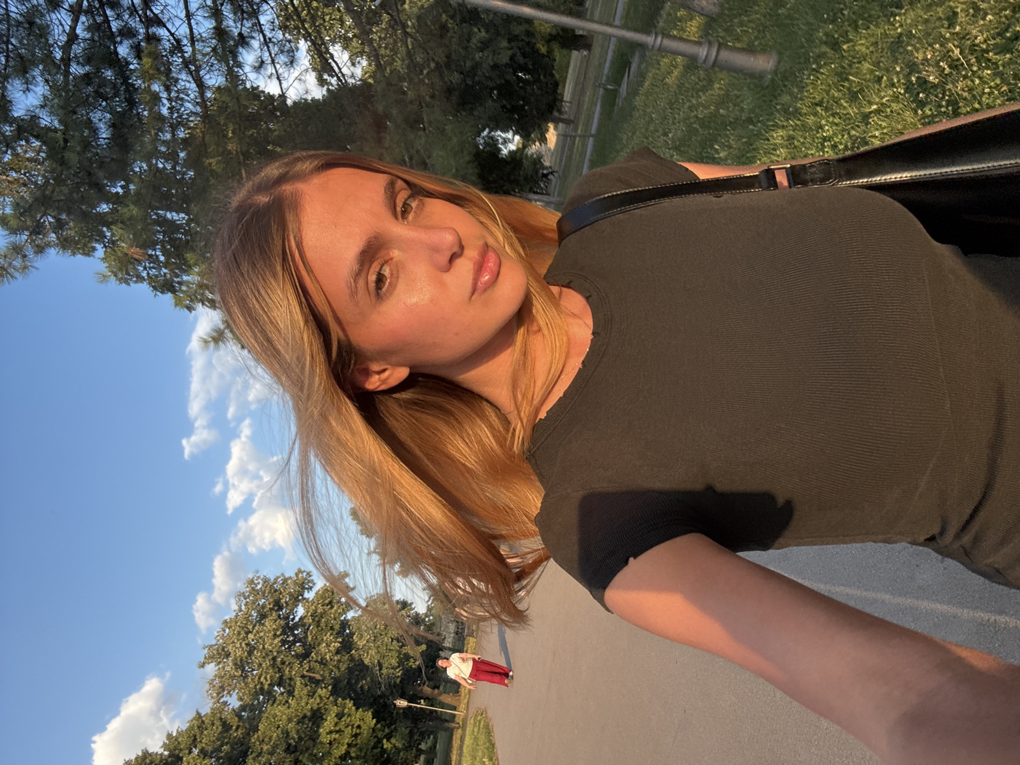 A young woman with long, blonde, wind-swept hair takes a selfie outdoors during golden hour. She is wearing a dark, ribbed t-shirt and a black shoulder bag. The background shows a paved pathway with a woman in red pants walking away from the camera, green grass, and trees. The sky is blue with white clouds. The lighting suggests it is late afternoon or early evening. This scene is likely in a park or a public walkway in Belgrade, Serbia, given the context.