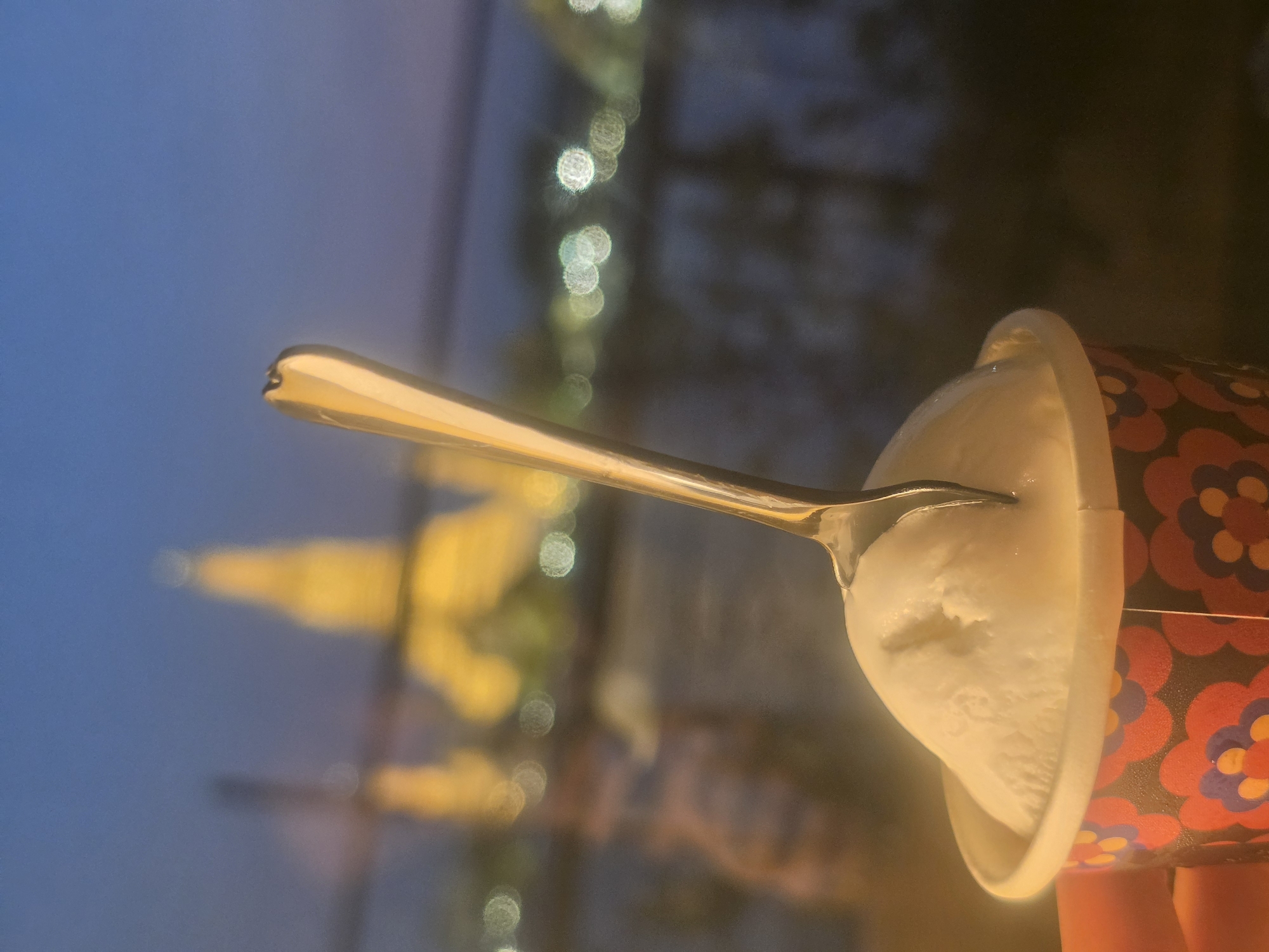 An image of a person with a container of white ice cream, pierced by a spoon, is presented in a blurry outdoor setting. The ice cream is in a paper cup with a red, brown, and blue floral pattern, and the spoon is silver in color. In the background, there are indistinct figures of buildings or structures, suggesting a possible urban environment. The image is taken in the city of Bangkok, Thailand, specifically near the Chao Phraya River, where the illuminated structure in the background might be Wat Arun (the Temple of Dawn). The overall lighting suggests an evening or nighttime setting.
