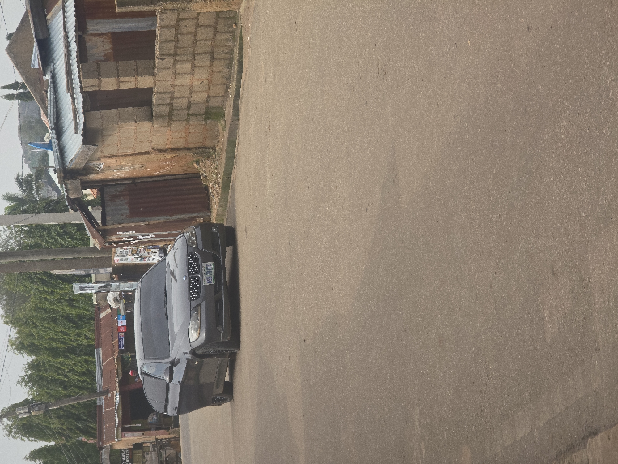 A dark gray BMW sedan with license plate ASB 791FC is parked on an asphalt road in Jos, Nigeria. The car is facing forward and slightly angled to the left. The driver's side door is open. To the left of the car, there is a building with a corrugated metal roof and signs. To the right of the car, there is a building constructed from concrete blocks with a corrugated metal roof. Trees line the background behind the buildings. The sky is overcast.