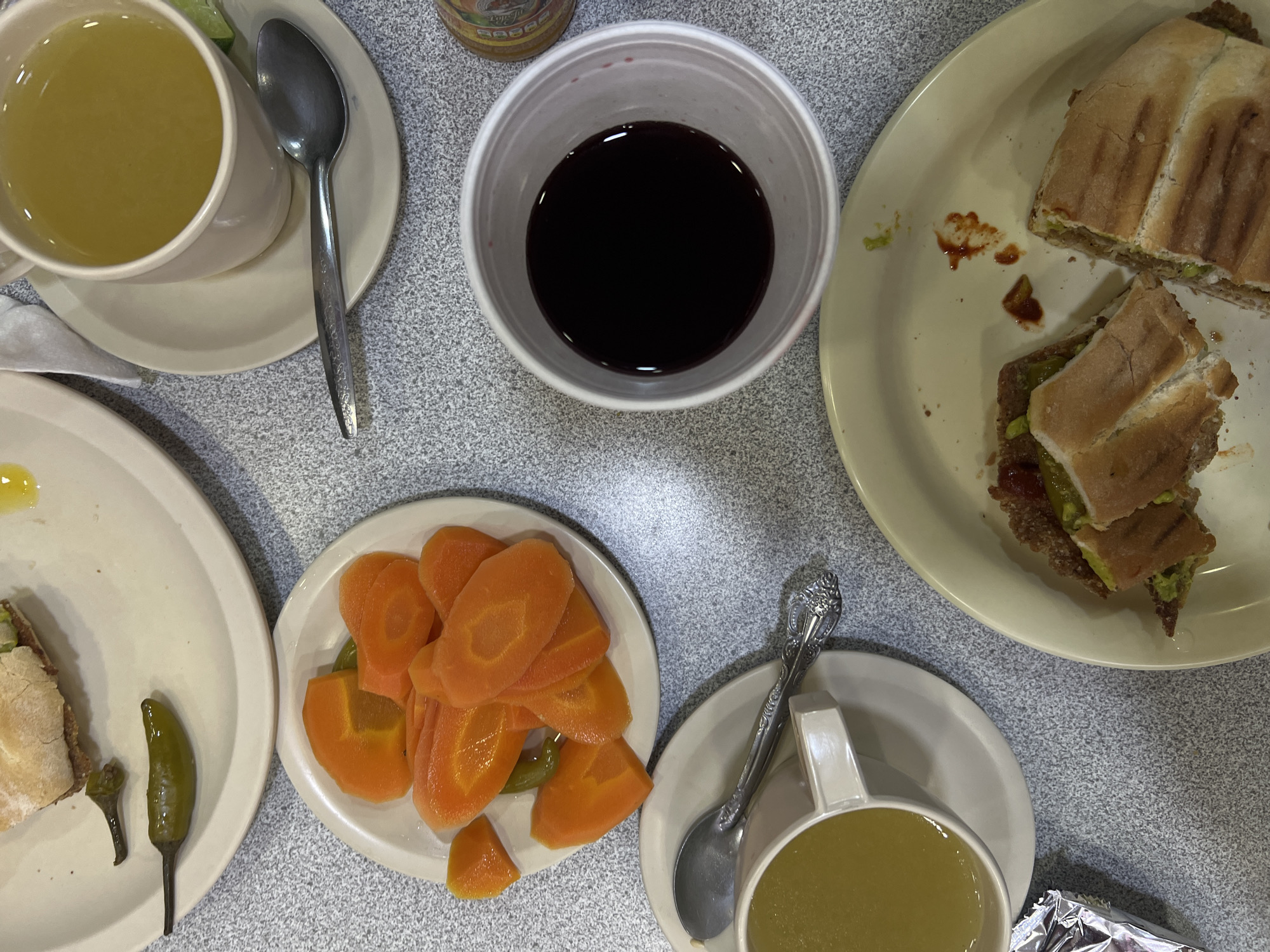 The image shows a table setting with a variety of food and beverages. There are two plates of sandwiches, one with pickled carrots and peppers, and two cups of broth. A cup of dark liquid, possibly coffee or tea, is also present. Utensils, including spoons, are scattered around the table. The setting appears to be a restaurant or cafe, based on the tableware and food presentation. While specific identifying details are limited, the presence of the Mexican hot sauce suggests that this is a casual dining establishment in Mexico City, Mexico.