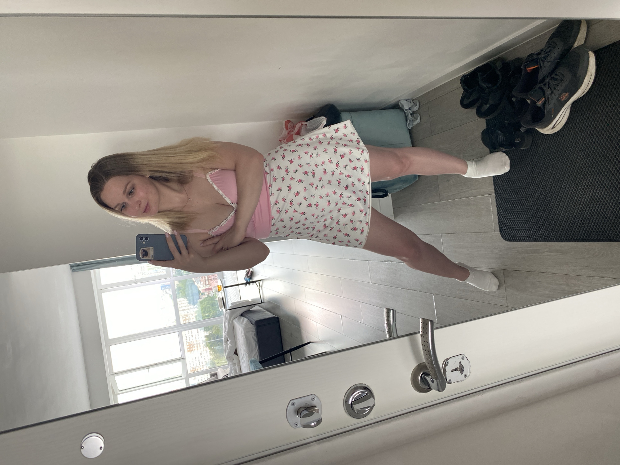 A young woman with blonde hair, wearing a pink top and a floral skirt, stands in front of a mirror. She is holding a smartphone in her hand and taking a selfie. The mirror reflects a bedroom with a bed, a window, and a door. The scene appears to be indoors. The woman's attire suggests a casual setting, possibly her home. The presence of a bed indicates a bedroom environment. The large window suggests the room might be on an upper floor, offering a view of the city. The overall atmosphere is relaxed and personal, captured in a moment of self-documentation.