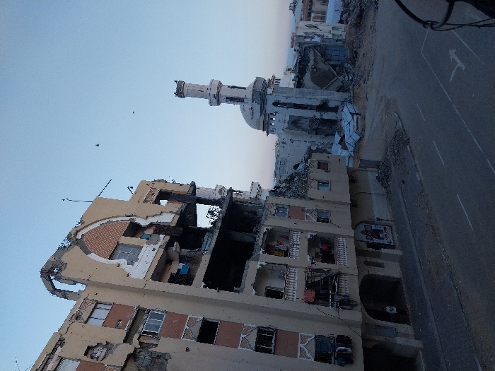 The image depicts a scene of extensive destruction in Khan Yunis, Palestinian Territory. The foreground is dominated by a multi-story residential or commercial building that has suffered severe damage. Large sections of its facade are torn away, revealing the interior structure, collapsed floors, and dark, burnt-out areas. Rubble and debris are scattered around and within the building.
In the mid-ground, a mosque with a distinct white minaret and dome is visible, also appearing to be damaged or partially destroyed, surrounded by debris. Further buildings in the background are likewise reduced to rubble or show significant structural damage.
A road or street runs through the scene, with a clear white arrow painted on its surface, indicating direction. The sky is a clear, pale blue, suggesting either late afternoon or early morning light, casting a somewhat somber mood over the devastation. No individuals are visible in the photograph. The scene conveys the profound impact of conflict or catastrophe. While some faint text might be present on a sign on the damaged foreground building, it is largely unreadable due to the damage and image quality.