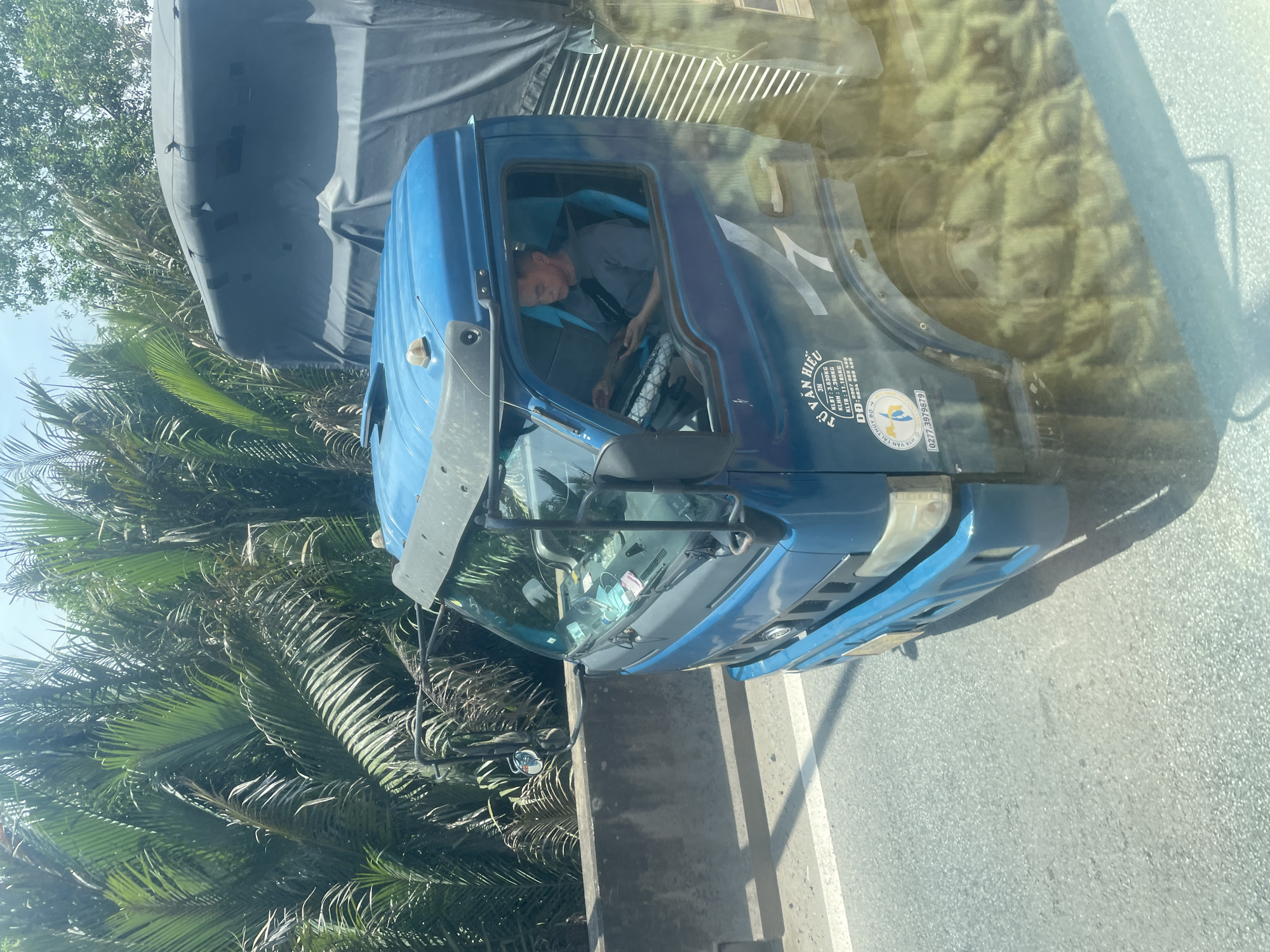 A blue truck is driving on a road in Dong Nai City, Vietnam. The truck has a black tarp covering its cargo. A man is driving the truck and looking at a tablet. The road is lined with palm trees and a concrete barrier. The sky is clear and sunny.