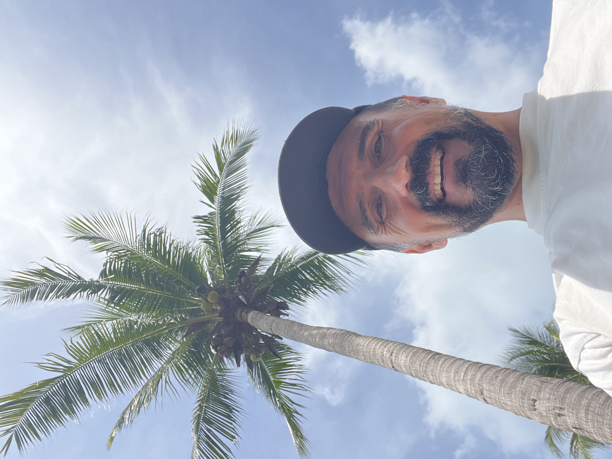 A man wearing a black baseball cap and a white t-shirt smiles broadly at the camera in a low-angle shot. He has a beard and dark hair. Behind him, a tall, textured palm tree trunk extends upwards, its fronds reaching towards a bright blue sky dotted with white clouds. Several green coconuts are visible clustered on the tree. The scene appears to be outdoors in a tropical location, consistent with Baan Chaweng Noi, Thailand, given the presence of the palm tree and clear sky.
