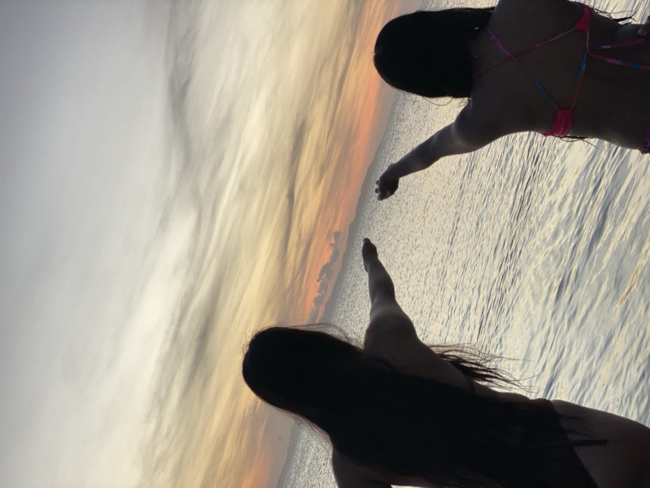 Two people, seen from behind, stand at the edge of water under a colorful sunset. Silhouetted against the vibrant sky, their outstretched arms reach toward each other. One wears a pink bikini. The background includes a mix of white and orange hues reflecting on the water, creating a serene and warm atmosphere.