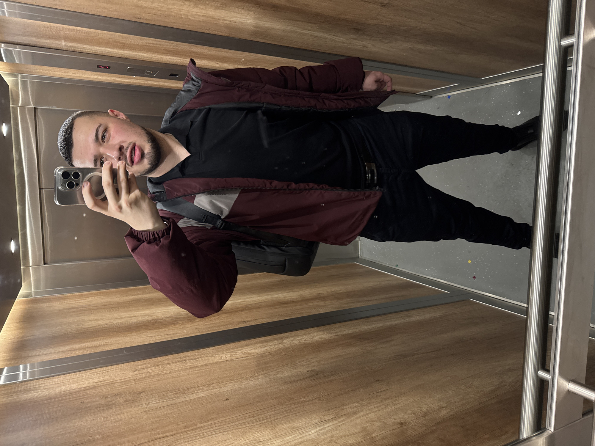 A man is taking a selfie in what appears to be an elevator. He is wearing a black t-shirt, black pants, and a burgundy and gray jacket. He has a backpack slung over his shoulder. The elevator walls are wood-paneled, and the floor is gray with confetti scattered on it. The location is unknown and cannot be determined from the image.