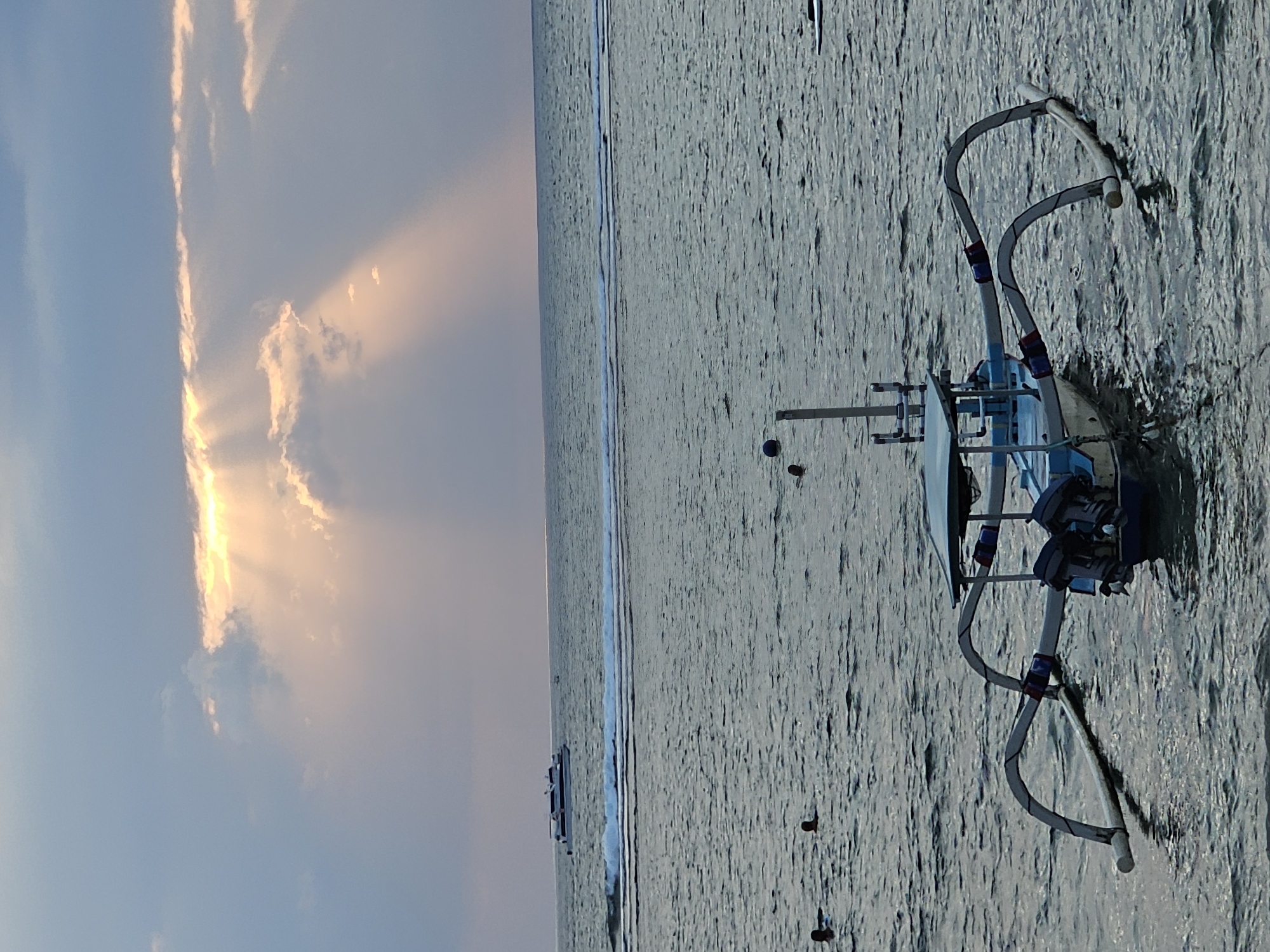 This is a photograph of a traditional Indonesian outrigger boat with a blue and white hull, two outboard motors, and a canopy, anchored in calm, dark water. In the distance, a larger white boat is visible on the horizon, with waves breaking near the shore. The sky is a mix of soft blues and grays, with dramatic sunbeams piercing through the clouds, suggesting either sunrise or sunset. Several people are swimming or wading in the water closer to the shore, their heads appearing as dark circles on the surface. The scene appears to be a coastal area, possibly a beach or harbor, in Denpasar, Indonesia.