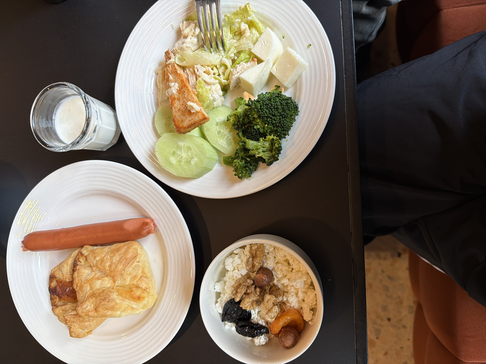 The image presents a tabletop view of a meal setup, seemingly within a domestic or dining setting, possibly in the city of Tbilisi, Georgia.
**Objects:**
* A plate with a salad consisting of lettuce, cucumber slices, broccoli florets, cheese cubes, and a piece of fish. A fork rests on the salad.
* A glass containing a white liquid, possibly milk or a similar beverage.
* A plate with a sausage and a pastry.
* A bowl filled with cottage cheese, nuts, and dried fruit.
**Subjects:**
* Partial view of a person wearing dark pants and sitting in a brown patterned chair is visible on the right side of the image.
**Environment:**
* The objects are set on a dark-colored surface that appears to be a table.
* The lighting is even, suggesting indoor illumination.
* The presence of multiple plates and a variety of foods indicate a meal is in progress.
The presence of the dried fruit, nuts, and cottage cheese, as well as the salad and sausage, suggests a combination of traditional and modern food options, potentially reflecting a blend of local and international influences often found in Tbilisi. The scene depicts a casual and personal dining experience.