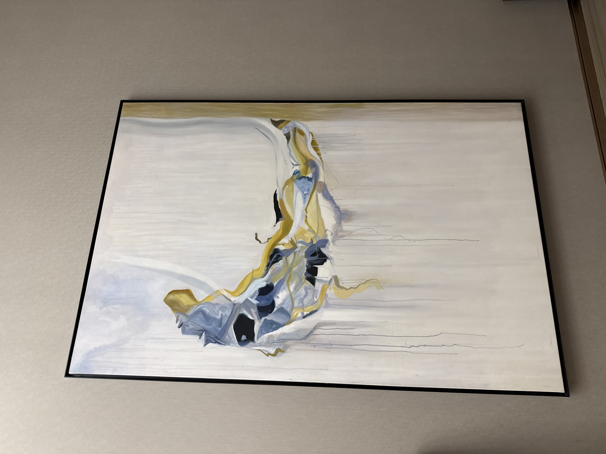 An abstract painting, presented in a black frame, is mounted on a light beige or off-white textured wall. The painting itself features a predominantly off-white background with subtle horizontal lines running across it. The central abstract element is a dynamic composition of flowing shapes and forms, primarily in shades of golden yellow, muted blue, grey, and deep black. These forms are intertwined, suggesting movement and fluidity, possibly resembling draped fabric or an organic, swirling mass. Some faint lines extend horizontally from this central cluster towards the right side of the canvas.
The wall exhibits a consistent, fine-grained texture. To the far right of the frame, a partial view of a light-colored door frame or wall molding is visible. The scene is indoors, likely a room or hallway. There are no people or discernible activities taking place, and no visible text is present within the image. The lighting is soft and even, highlighting the artwork. The location context is Khimki, Russia.