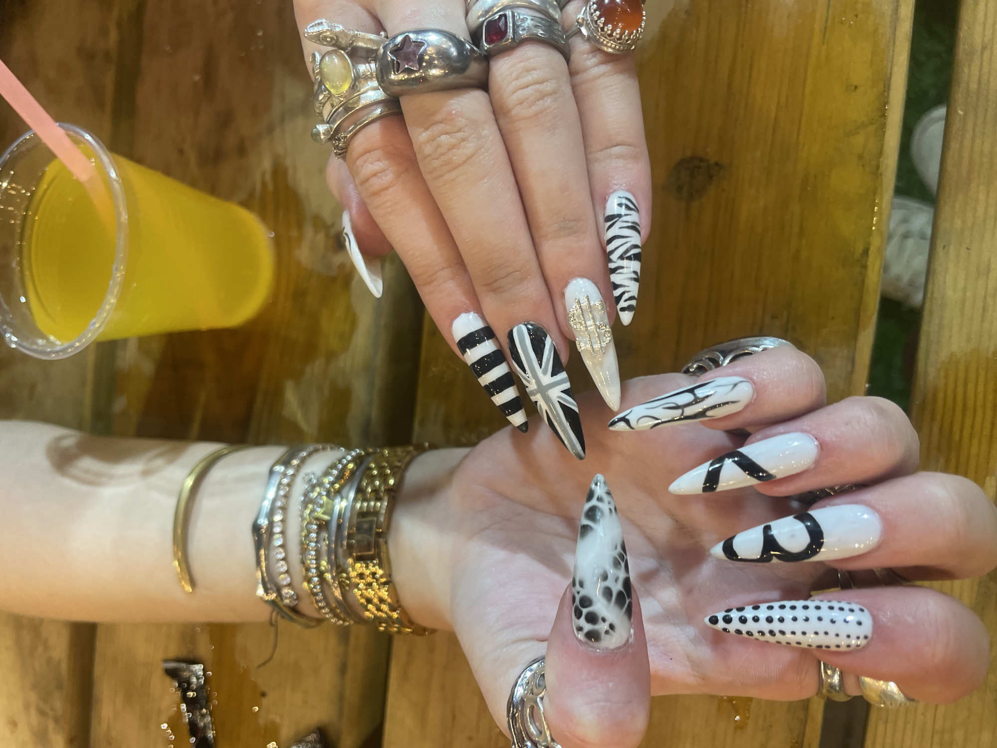 A photograph depicts two hands with long, stiletto-shaped nails adorned with various black and white patterns and embellishments. The fingers of one hand are adorned with multiple silver rings featuring various gemstones. The arm of this hand is decorated with several bracelets. Next to this hand is a clear plastic cup filled with a yellow liquid and a pink straw. The background appears to be a wooden surface. The location is Raanana, Israel.