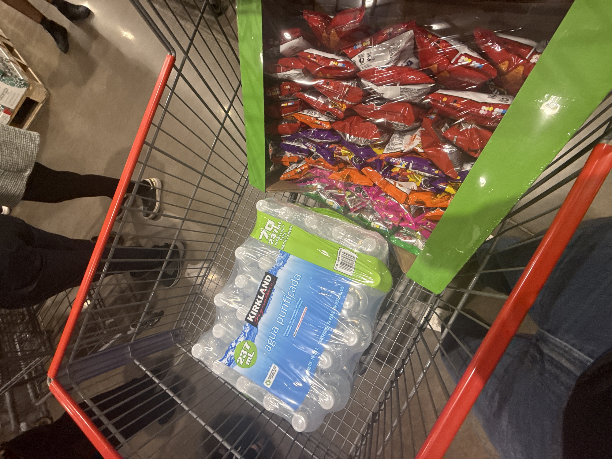 An overhead view captures the interior of a large retail store, likely a warehouse club such as Costco in Puebla, Mexico. A red-handled shopping cart is prominently featured, filled with bulk items. At the bottom of the cart, a shrink-wrapped pack of 70 small bottles of Kirkland Signature purified water is visible. The label specifies "agua purificada," "237 mL," and "CONTENIDO 70 PZAS DE 237 mL C/U," with a total net volume of "16.5 L," and directs visitors to "www.costco.com.mx."
Stacked above the water bottles is a large open box containing numerous individual bags of assorted snacks, including chips and candies in various red, purple, orange, and pink packaging.
Parts of at least three individuals are visible around the cart. On the left, a person wearing dark pants and shoes stands near a wooden pallet, alongside another person in a grey sweater, black pants, and white shoes. On the right, a person in blue jeans and dark shoes appears to be pushing the cart. The store's floor is concrete, typical of warehouse-style retailers. The bright artificial lighting suggests an active shopping environment.