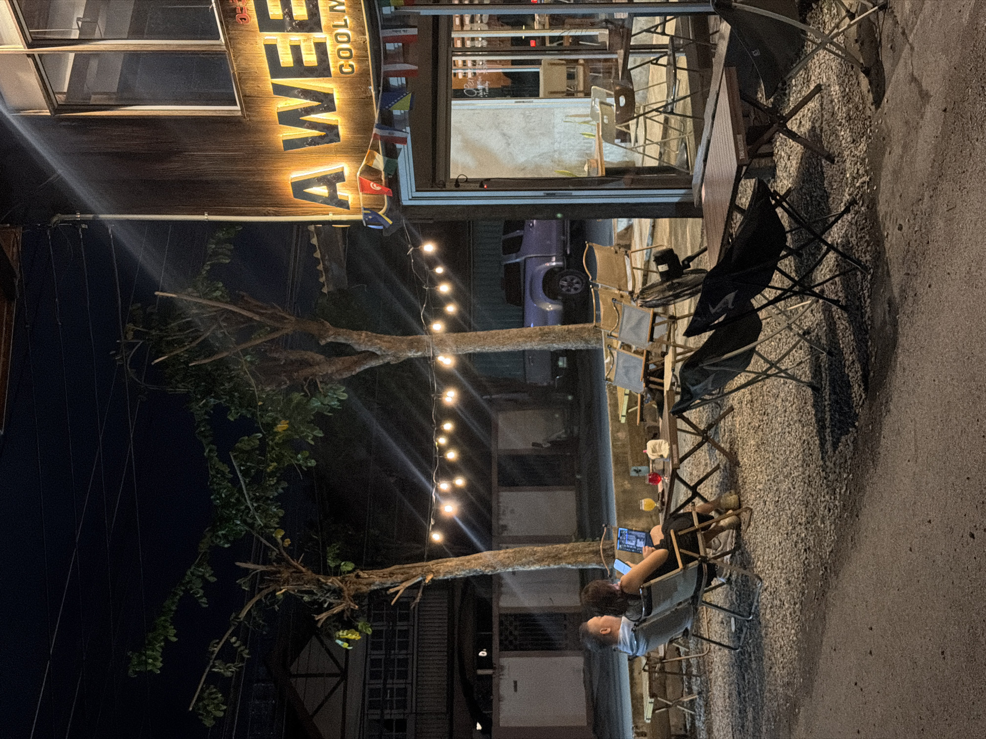 The image depicts an outdoor seating area of a cafe at night, likely in Chiang Mai City Municipality, Thailand. A couple is seated at a small table in the foreground, with the woman looking at her phone and a laptop open in front of her. Two drinks, one orange and one red, are also on the table. In the background, a sign with illuminated letters reads "A WEEKEND." The cafe has a rustic wooden exterior and is adorned with a string of lights and small flags from various countries. Several empty chairs and tables are arranged on a gravel surface. The scene is illuminated by artificial lights, creating a warm and inviting atmosphere.