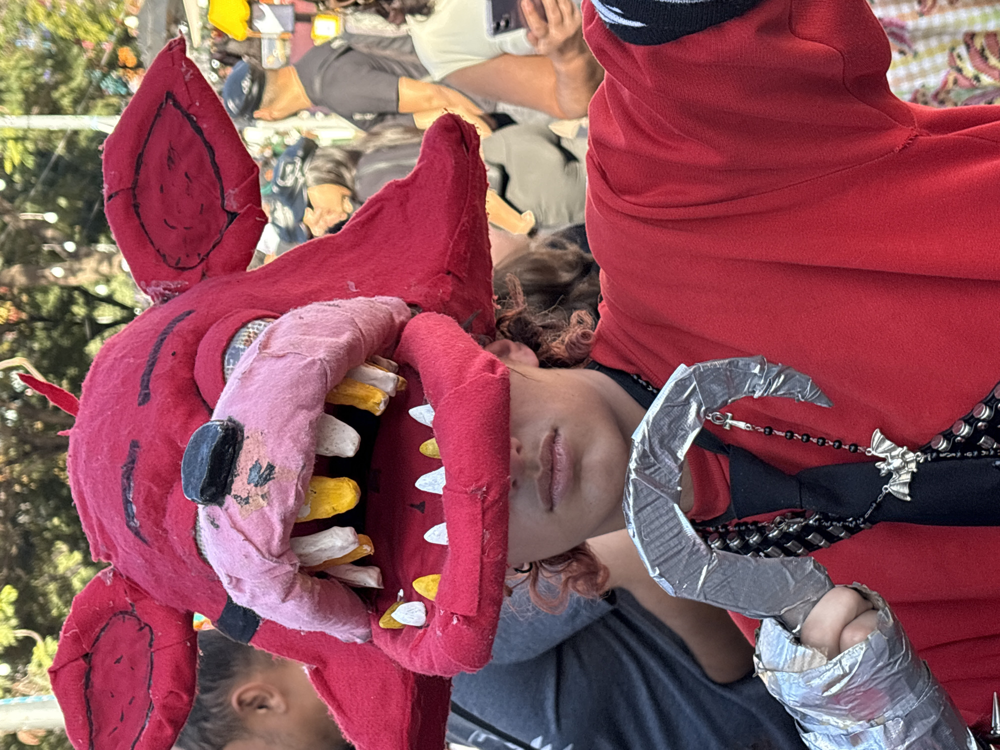The image shows a person in costume at an outdoor event. The person is wearing a large, red, plush fox head mask that covers their face and extends to their shoulders. The mask has large ears, black-outlined eyes, a pink snout, and a wide mouth with prominent, uneven teeth that are white and yellow. The mouth is partially open, revealing the teeth. The person is wearing a red shirt and a black tie with a beaded necklace that has a bat charm. On their left arm, they are wearing a silver, metallic-looking gauntlet. In the background, other people are visible, some wearing hats and casual clothing. The environment appears to be a street or plaza with trees and what look like market stalls or food vendors. The lighting suggests it is daytime. The scene is likely a public gathering, possibly a festival or convention, in the city of São Paulo, Brazil.