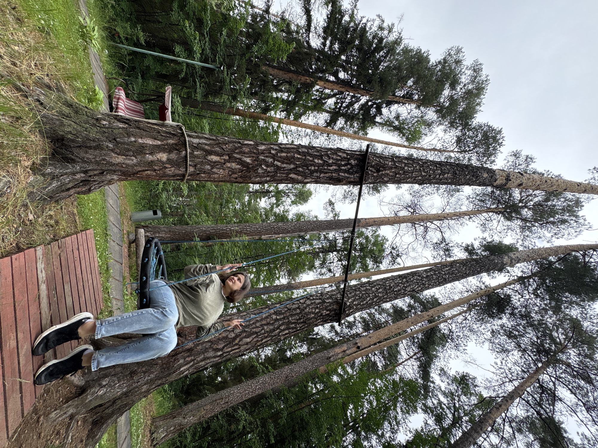 A young woman with short, dark hair is sitting on a rope swing in a forest. She is wearing a green long-sleeved shirt, blue jeans, and black sneakers. The swing is suspended between two tall pine trees. The ground is covered with grass and fallen leaves. In the background, there is a red and white striped swing set and a trash can. The sky is overcast. The location is the village of Kostovaty, Russia.