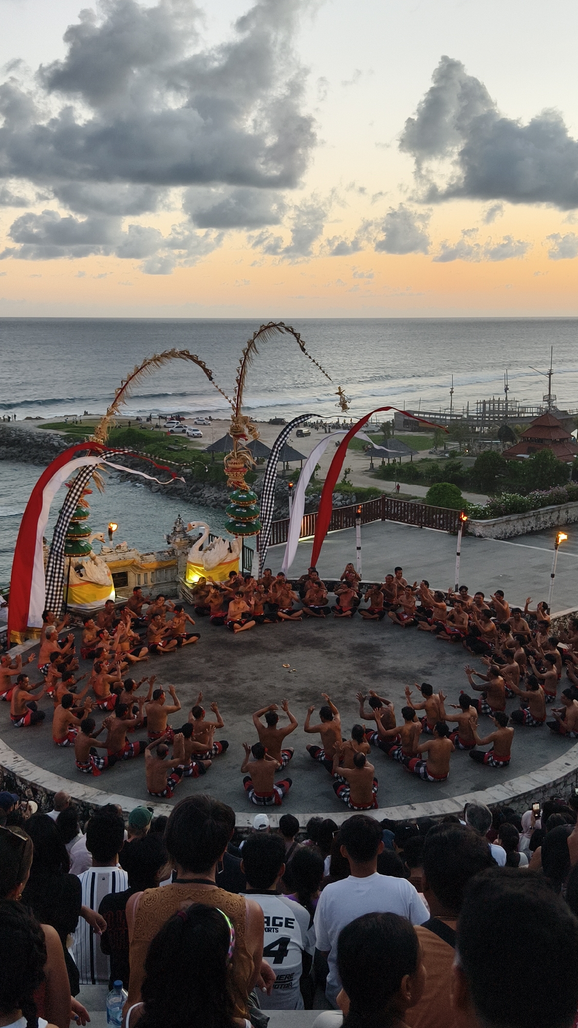 The image shows a performance of the Kecak dance in Kuta Selatan, Indonesia. A large group of shirtless men wearing patterned cloths around their waists are seated in a circle on a circular stage. They are raising their hands in unison as part of the dance. Behind the stage are decorated structures, including curved bamboo poles and flags. Beyond that is a view of the ocean at dusk, with a breakwater and some buildings visible. In the foreground, a crowd of spectators is watching the performance.