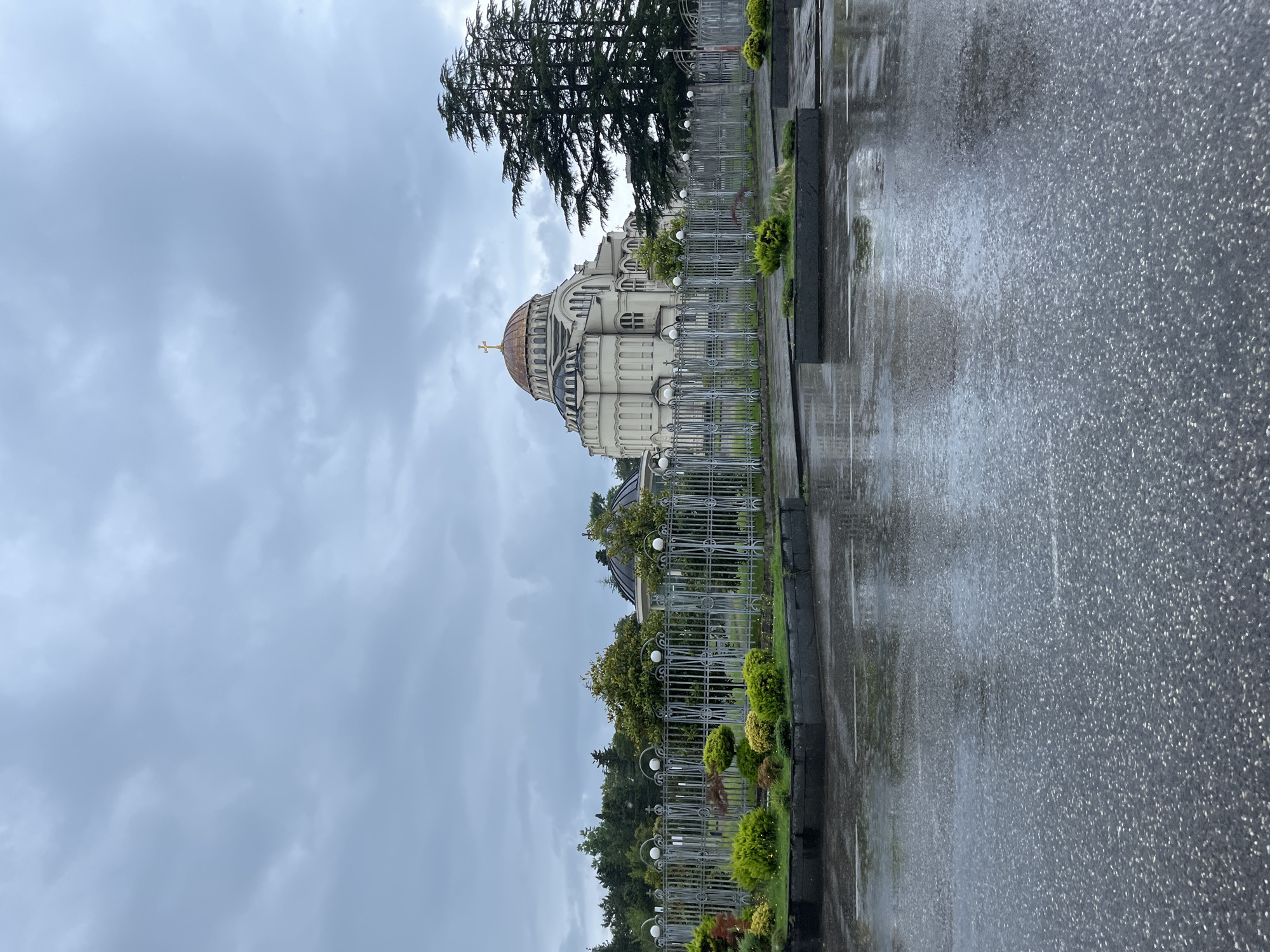 The image shows the Holy Trinity Cathedral in Poti, Georgia, on a cloudy and rainy day. The wet asphalt in the foreground reflects the overcast sky and the surrounding landscape. A decorative metal fence encloses a garden area with manicured shrubs and small trees. Beyond the fence, the large, imposing structure of the Holy Trinity Cathedral dominates the scene. It is characterized by its distinctive dome topped with a golden cross. The cathedral's architecture appears to be neoclassical with Byzantine influences. Lush green trees, including a tall evergreen tree on the right, frame the building. The overall atmosphere is somber, likely due to the weather. There are no people or animals visible in the frame.