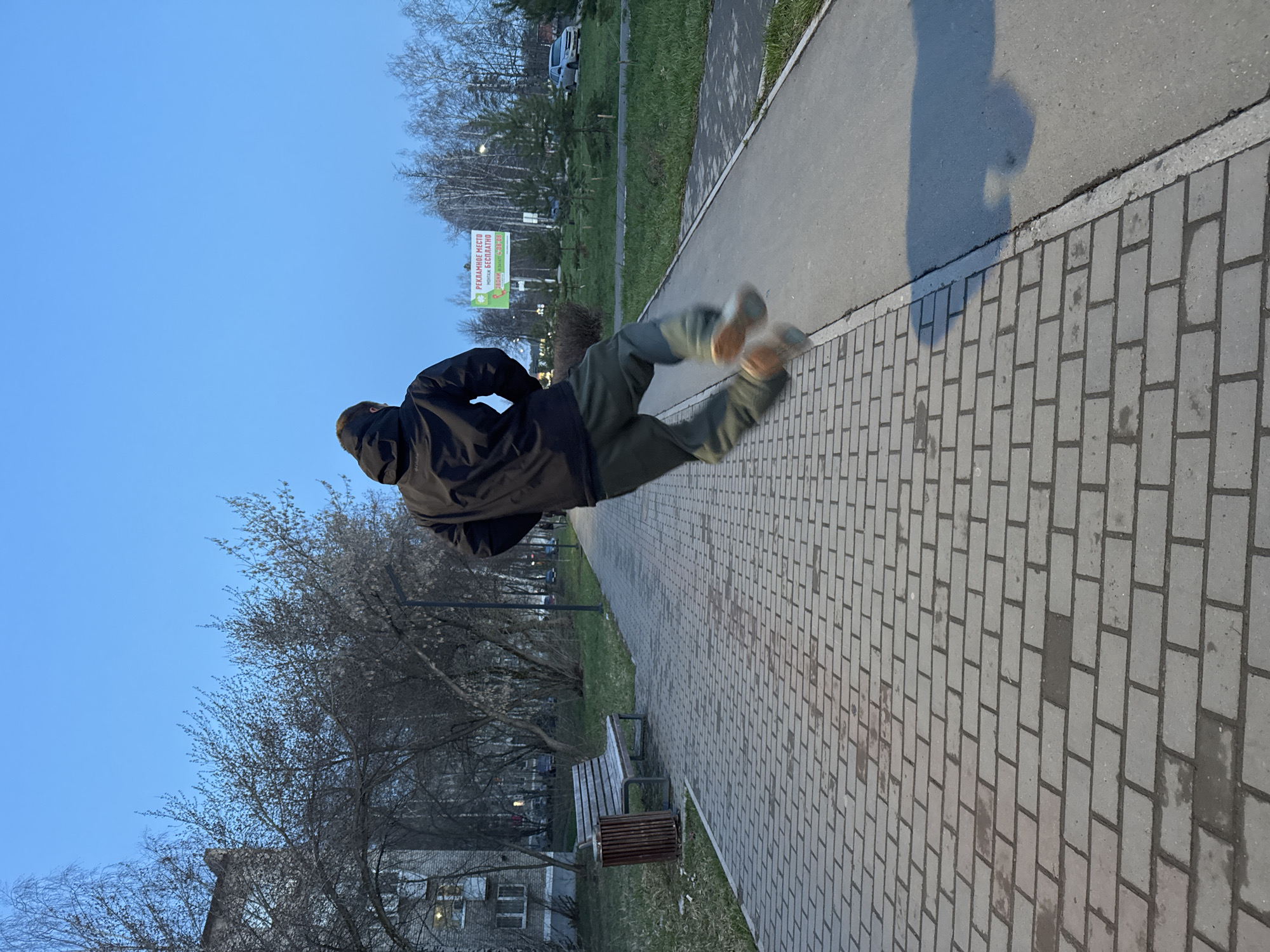 An outdoor scene is depicted, rotated 90 degrees clockwise, giving the illusion of a person floating horizontally mid-air above a paved pathway. The person, seen from behind, wears a dark hooded jacket, olive green or dark trousers, and light-colored athletic shoes. Their body is arched, suggesting a dynamic jump or levitation.
The ground comprises a light grey paved path made of rectangular bricks, running alongside a darker asphalt path. To the left of the paved path (when viewed upright), there is a strip of green grass, transitioning to numerous bare-branched trees, some showing early budding leaves, characteristic of late winter or early spring. In the background, partially obscured by trees, are multi-story residential buildings and a distant light-colored car. A wooden slat bench and a cylindrical slatted trash bin are positioned beside the paved path.
A distinct, elongated shadow of a person, likely the photographer or another individual, is cast across the asphalt path, indicating a low sun angle. The sky above is clear and bright blue.
A white rectangular sign with a green border and a green leaf logo is visible in the middle ground. The Cyrillic text on the sign reads:
- In red: "РЕКЛАМНОЕ МЕСТО" (REKLAMNOYE MESTO), meaning "Advertising Space."
- In green: "УСПЕЙ ЗАНЯТЬ БЕСПЛАТНО!" (USPEY ZANYAT' BESPLATNO!), translating to "Hurry to occupy for FREE!"
- In smaller red text: "ЗВОНИ:" (ZVONI:), meaning "Call:", followed by a partially visible phone number.
The lighting suggests late afternoon or early evening. The scene takes place in an urban or suburban park environment, consistent with Chaikovsky, Russia.