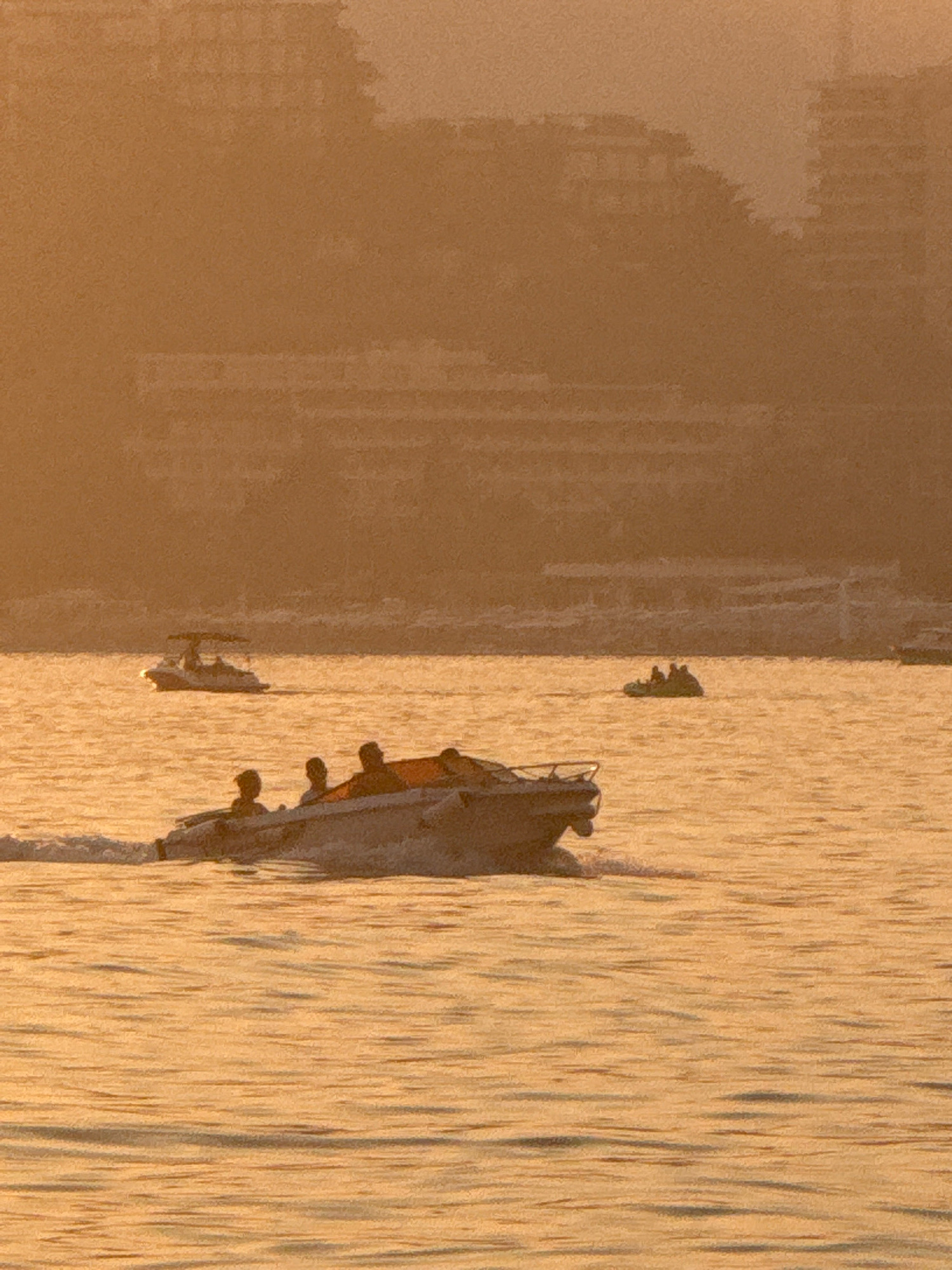A scenic, golden hour photograph captures a body of water, likely the Adriatic Sea, with several boats visible. The water's surface reflects the warm, diffused sunlight, creating a textured, shimmering effect. In the foreground, a speed boat is actively moving, its wake trailing behind. Several people are seated in this boat. Further in the distance, another, smaller boat with at least two individuals can be seen. In the background, hazy, silhouetted structures, likely buildings along the coast of Bečići, Montenegro, rise against the golden sky, suggesting a coastal city setting. The overall impression is one of a tranquil, late afternoon or early evening on the water.