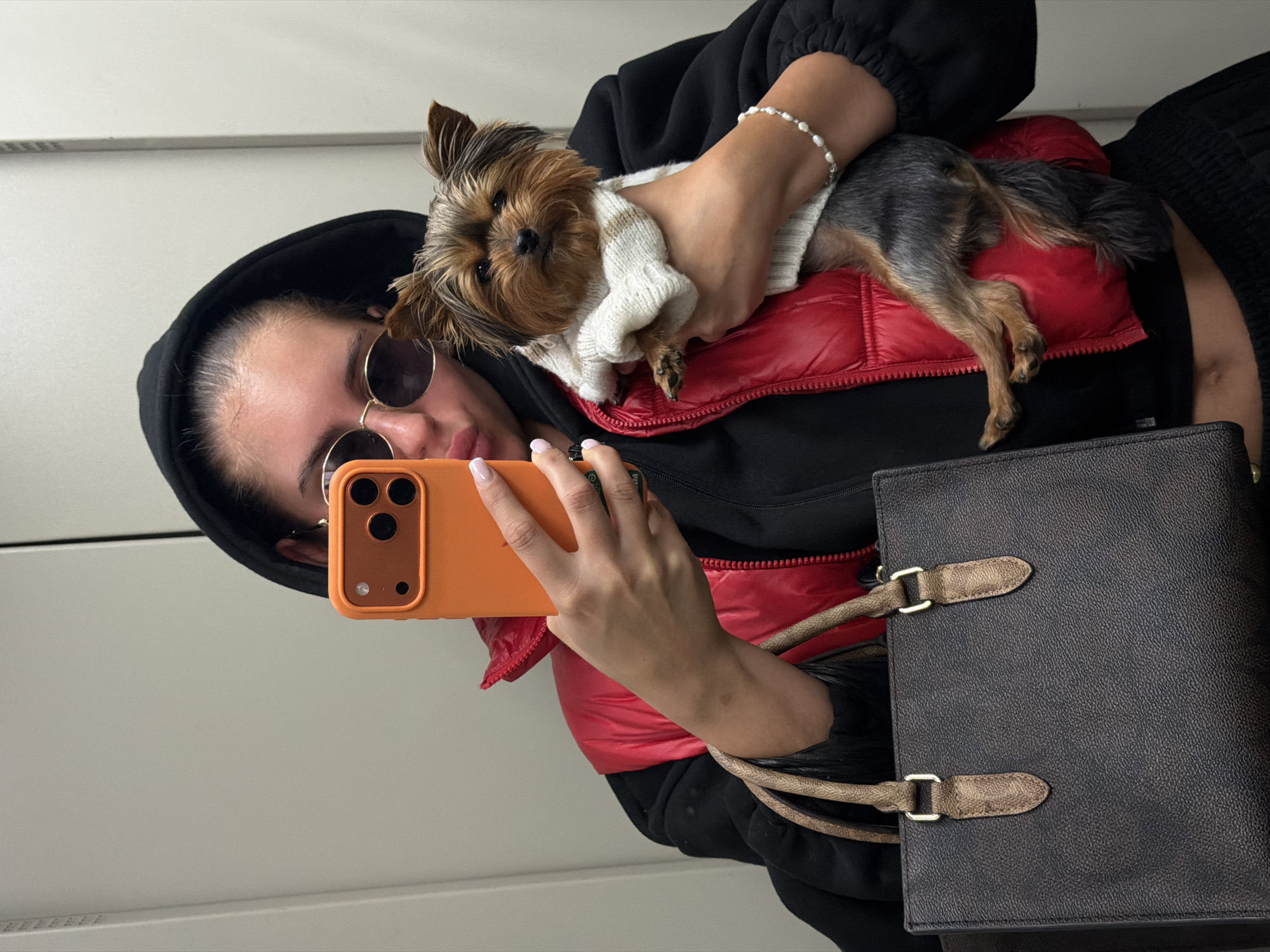 A woman with sunglasses and a black hooded jacket is taking a selfie in front of a white or light-colored wall. She is holding a Yorkshire terrier puppy wearing a cream and brown sweater. The woman also wears a red puffer vest and a pearl bracelet on her left wrist. She holds an orange smartphone with her right hand. A brown leather purse with tan straps is visible on the right side of the frame. The location, Divnogorsk, Russia, cannot be determined from the image.
