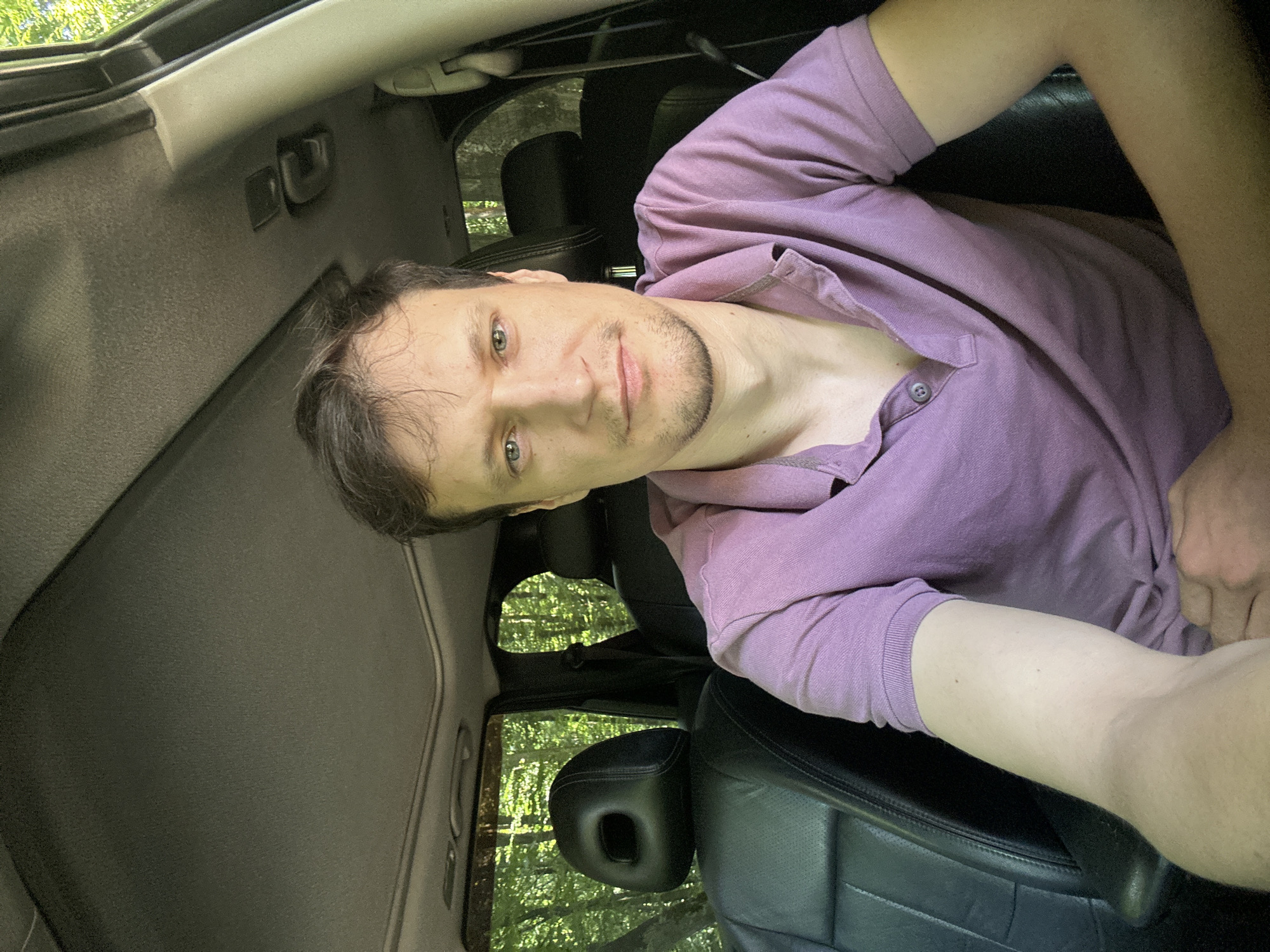 A man is seated inside a vehicle, likely a car, with a panoramic sunroof overhead. The interior of the car appears to be black leather. Outside the vehicle, a dense forest is visible through the windows, suggesting the car is parked in a wooded area. The man is wearing a purple polo shirt. His pose suggests he is taking a selfie. The background appears to be a natural, outdoor environment. There are no other individuals or significant objects visible in the immediate vicinity of the man inside the car, other than the car's interior elements. The lighting suggests it is daytime, with sunlight filtering through the trees. The provided context that the location is Shatava, Ukraine, indicates the setting is likely a rural or natural area in that region.