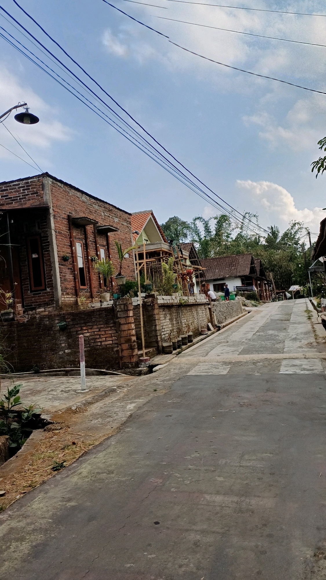 The image shows a street scene in Tengahan, Indonesia. The street is paved and slopes slightly uphill. On the left side of the street, there is a brick building with wooden window frames and potted plants. A street lamp is visible above the building. Further up the street, on the right, are several houses with traditional Indonesian architecture, featuring wooden structures and tiled roofs. Some people are visible near the houses. The background consists of lush green trees and a cloudy sky. The overall impression is a rural or semi-rural residential area in Indonesia.