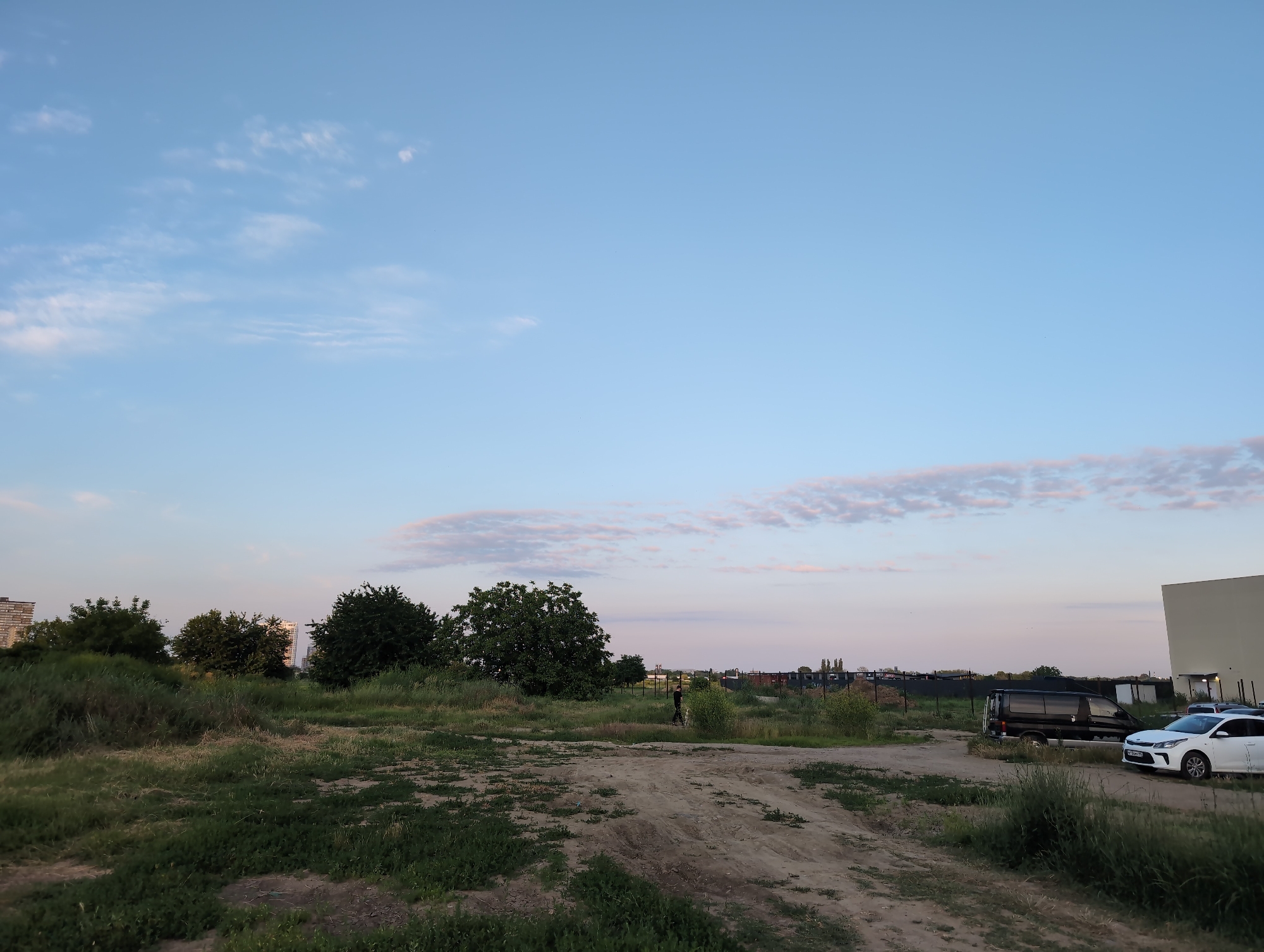 The image depicts an outdoor scene at dusk in Krasnodar, Russia. A dirt road cuts through a grassy field with scattered trees. In the distance, several buildings and a fence are visible. A person is walking with a dog on the road, and a black van and a white car are parked near a building on the right side of the frame. The sky is clear with some wispy clouds, and the light suggests it is either early morning or late evening. The overall atmosphere is calm and serene, with the natural landscape contrasting with the man-made structures.