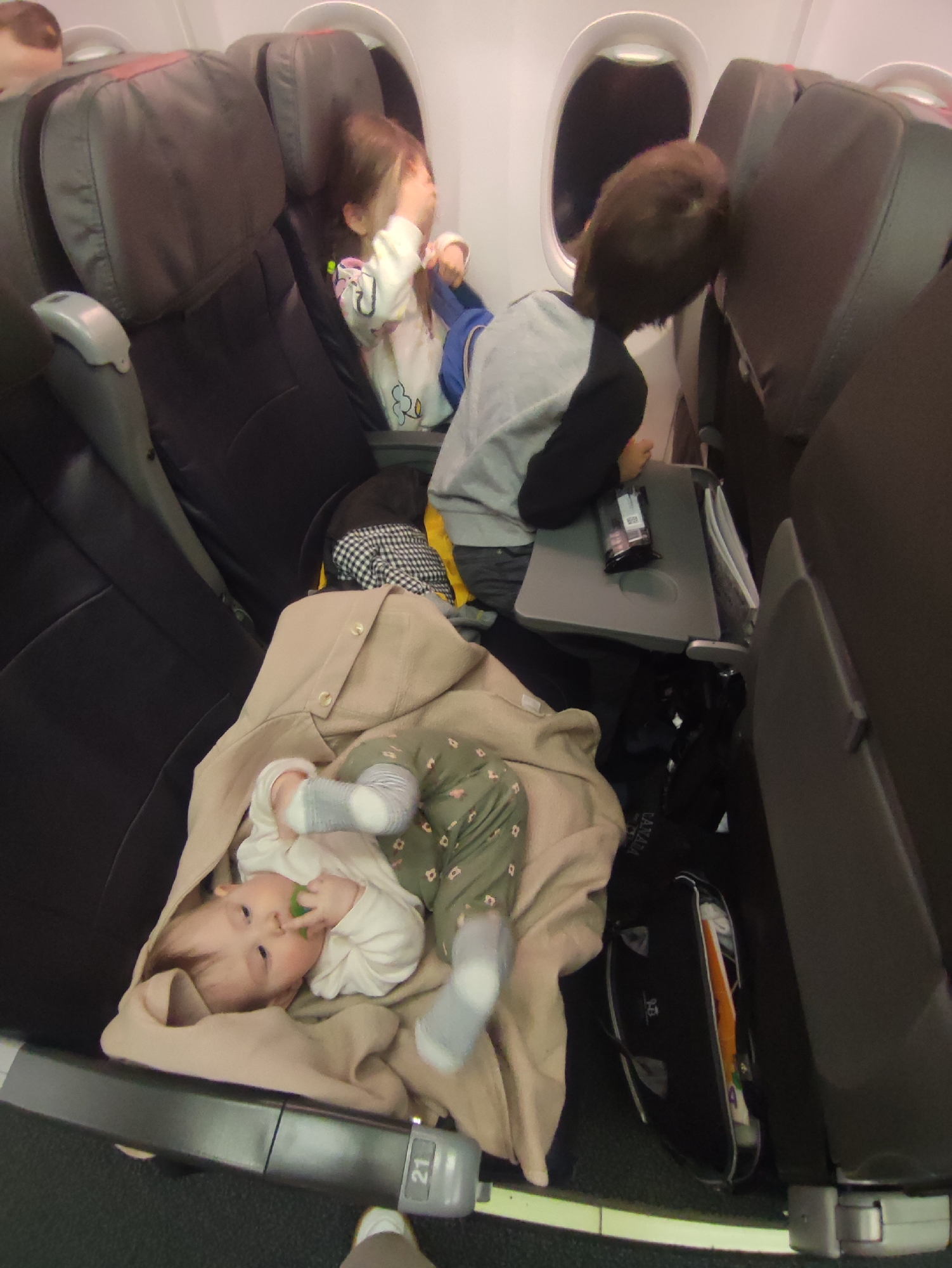 The image depicts the interior of an airplane cabin, specifically observed in the context of the city of Sochi, Russia. The scene captures four individuals: three children and a partial view of an adult. In the foreground, a baby lies on its back, partially swaddled in a tan garment, wearing a white top and patterned green pants with grey-and-white socks. The baby is looking towards the camera, holding a green object in its mouth. An armrest with the number "21" is visible in front of the baby. In the middle ground, in the row ahead, a young girl with brown hair is seated by an oval window, wearing a light-colored shirt, and appears to be rubbing her face with her right hand. Next to her, an older child, identified as a boy with brown hair and wearing a grey and black shirt, leans forward over a deployed tray table. A dark package and documents are on the tray table. A black bag, partially revealing items with orange and white colors and bearing a "CANADA" tag, rests on the floor beneath the tray table. Further back and to the left, a blurry partial view of an adult's head is discernible. The seats are upholstered in a dark material with lighter grey armrests. The floor is carpeted in a dark tone.