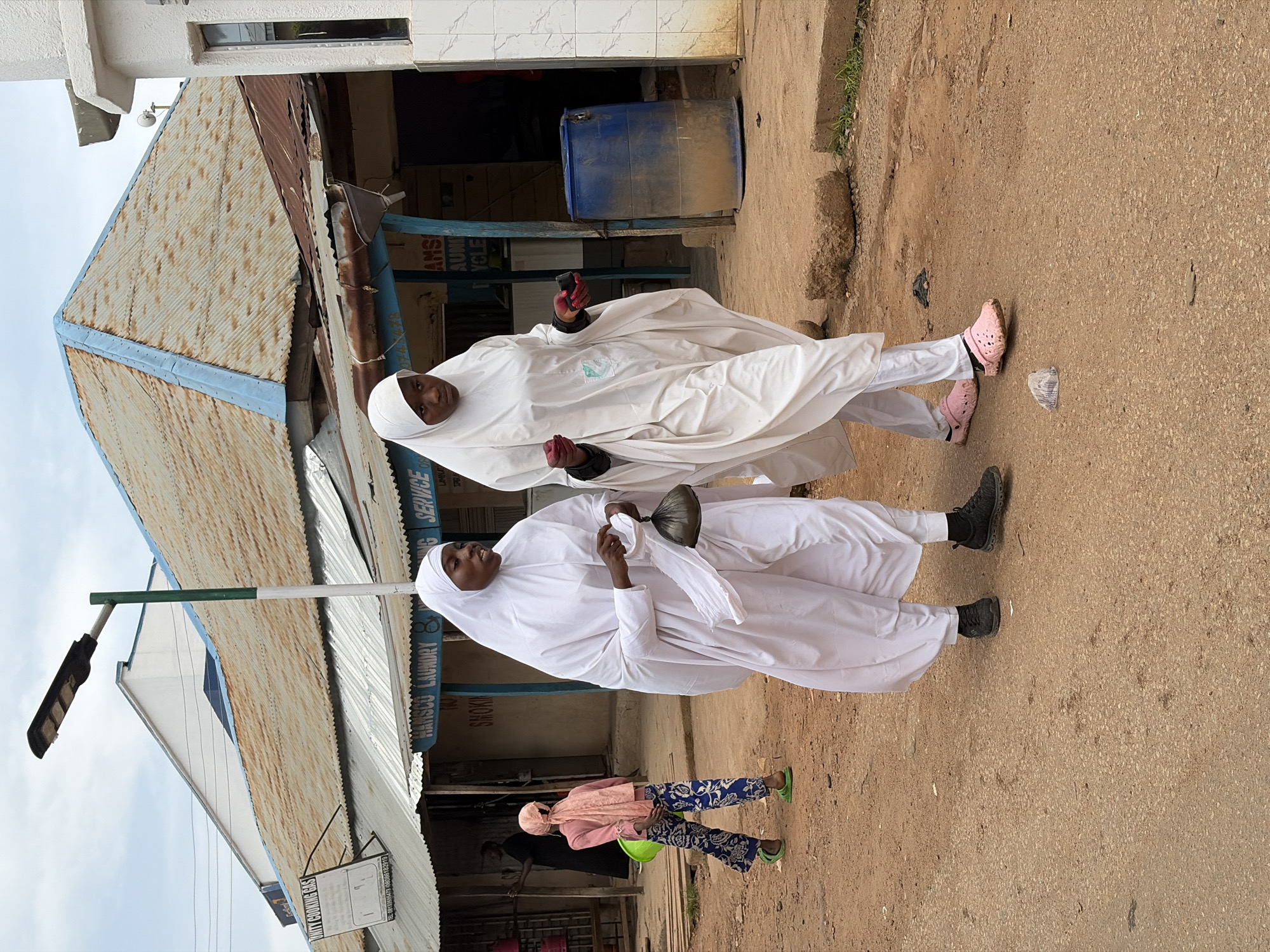 Two women wearing white hijabs and long white garments walk on a dirt road in New Stadium, Nigeria. The woman on the left is wearing black sneakers, and the woman on the right is wearing pink Crocs. Both are walking towards the right side of the image. In the background, there are several buildings with corrugated metal roofs. One building has a sign that reads "HANSCU LAUNDRY" and another sign that reads "SERVICE." A blue plastic barrel is visible next to a building on the right side of the image. A young child in a pink headscarf and blue patterned pants is walking away from the camera to the left, carrying a green bucket. The sky is overcast.