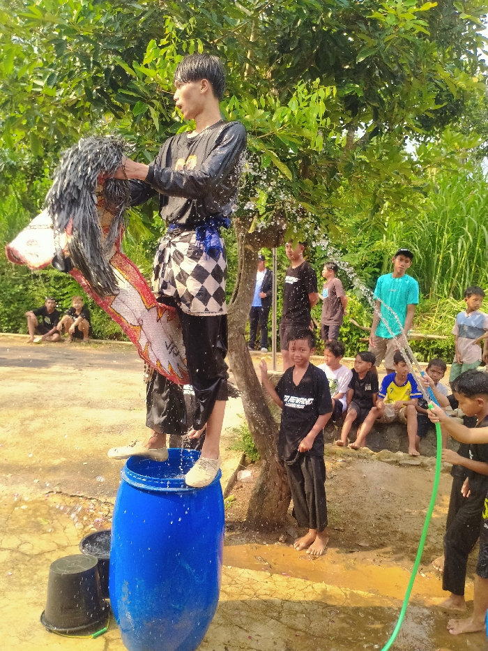 The image depicts an outdoor scene in Gebyog, Indonesia, capturing an activity involving a central figure and several onlookers. A young adult male, visibly wet, stands balanced atop a blue plastic barrel in the foreground. He wears a black long-sleeved shirt, black pants, a black-and-white checkered sarong tied at the waist, and light-colored shoes. He holds a decorated, traditional horse-head puppet, likely a Kuda Lumping prop, which is also wet and features a prominent black mane. Water actively splashes from his person and the puppet.
To the immediate right of the central figure, a young boy, barefoot, wearing a wet black t-shirt and dark pants, stands looking towards the camera with a slight smile. Further right, another young boy, also wet, operates a green hose, directing a stream of water into the scene.
In the background, a group of approximately eight to ten boys and young men observe the activity. Some are seated on a low, irregular stone or concrete ledge, while others stand behind them. Two black plastic buckets are visible on the ground near the base of the blue barrel. The ground beneath the participants is damp, exposed earth, with a drier, possibly paved, path visible further back where some observers stand. The environment is characterized by lush green foliage, including trees and tall grasses, which form a dense backdrop under bright daylight conditions.