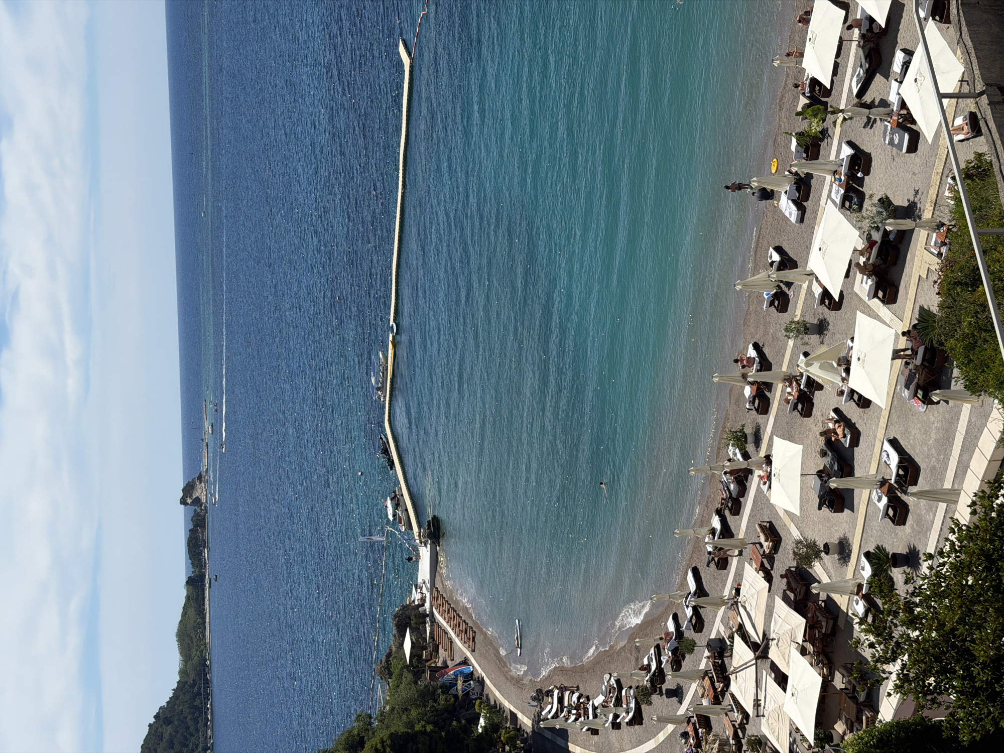 This image captures a panoramic view of a beach in Budva, Montenegro, likely the Mogren Beach area, on a sunny day. The turquoise waters of the Adriatic Sea stretch to the horizon, meeting a bright blue sky dotted with white clouds. A long, floating pontoon stretches out into the sea, serving as a walkway or a diving platform.
The foreground and midground of the image are dominated by a beach set up with numerous lounge chairs and white umbrellas, indicating a resort or a well-maintained public beach. Several people are visible on the beach, either relaxing on the lounge chairs, sunbathing, or wading in the shallow waters. One person is seen swimming further out in the sea, and another is standing near the water's edge. A paddleboard is floating near the shore. Boats are visible in the distance, both moored and in motion, and a small rocky island with vegetation is situated offshore. The beach itself appears to be a mix of sand and pebbles. Lush green trees and vegetation cover the hillside bordering the beach, adding to the scenic beauty of the location.