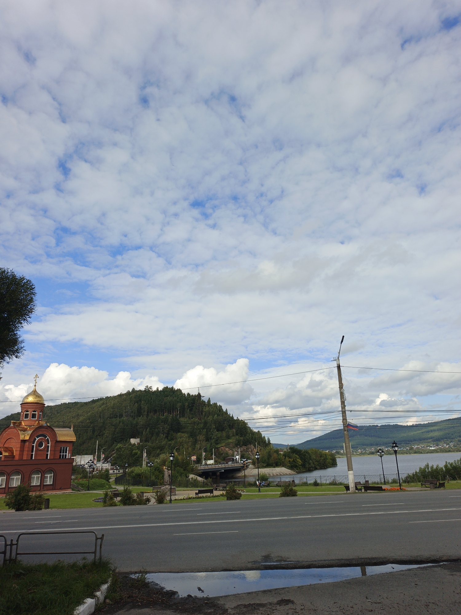 A wide-angle, low-angle exterior shot captures a bright, cloudy day scene in Zlatoust, Russia. In the foreground, a wet asphalt road stretches across the bottom of the frame, with a puddle reflecting the sky. A low metal barrier runs along the edge of the road on the left. To the left, a red brick building with a golden dome, resembling a church, is visible. White arched windows are present on the facade of the building. Behind the building and to the right, a lush, tree-covered hill dominates the middle ground. A body of water, likely a river, lies beyond the hill, with a distant city and more rolling hills visible on the far side. Several lampposts and benches are scattered along the waterfront. A bridge spans the water in the distance. Overhead, a vast expanse of blue sky is filled with fluffy white clouds. The overall impression is a serene landscape, with a prominent architectural element in the foreground.