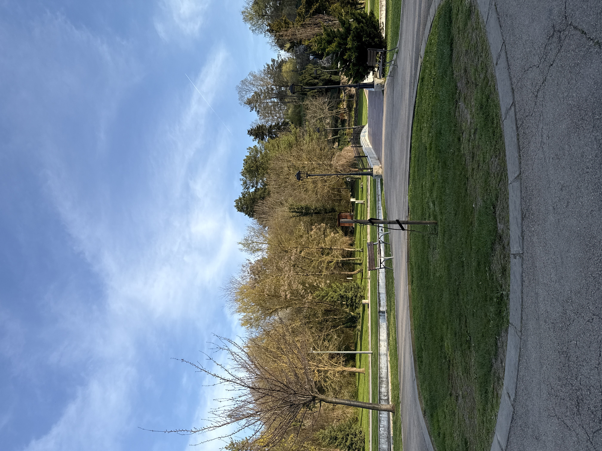 The image shows a park in Dobrich, Bulgaria. The scene captures a paved path curving along a circular grassy area. Along the path, there are several benches and lampposts. Deciduous trees with budding leaves line the park, and a bare tree stands prominently in the foreground. The sky is blue with scattered white clouds and a contrail is visible.
