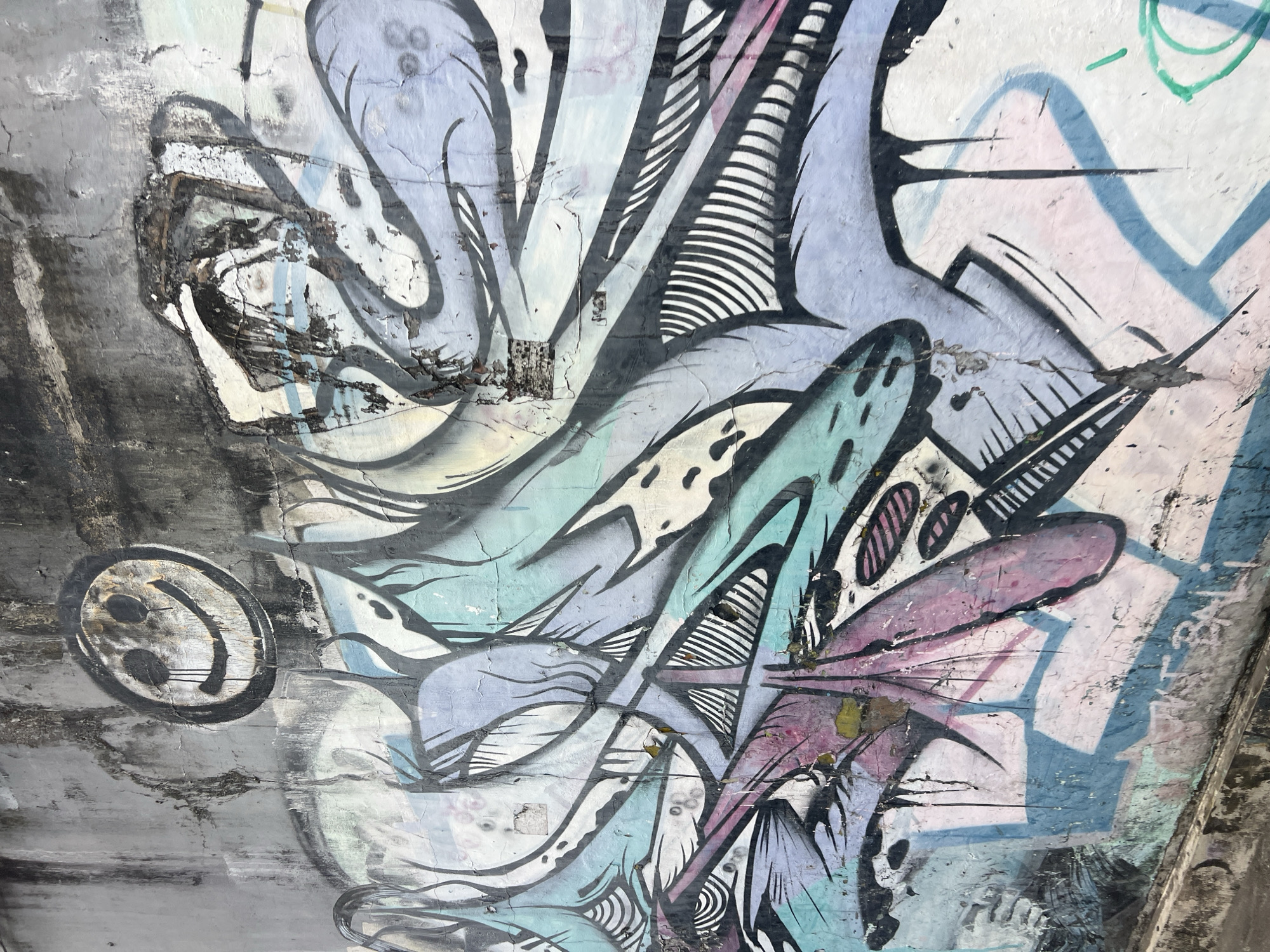 The image shows a section of a concrete wall in Kuta, Indonesia, covered with graffiti. The graffiti features abstract shapes in shades of blue, purple, teal, and black, with some areas of white and pink. There is a stylized smiley face at the top left, appearing to be crying. The wall itself is weathered and cracked, with visible signs of age and decay, including peeling paint and dirt streaks. The lower part of the image shows the base of the wall, which appears to be a concrete curb or ledge, also showing wear. The overall impression is of an urban environment with street art.
