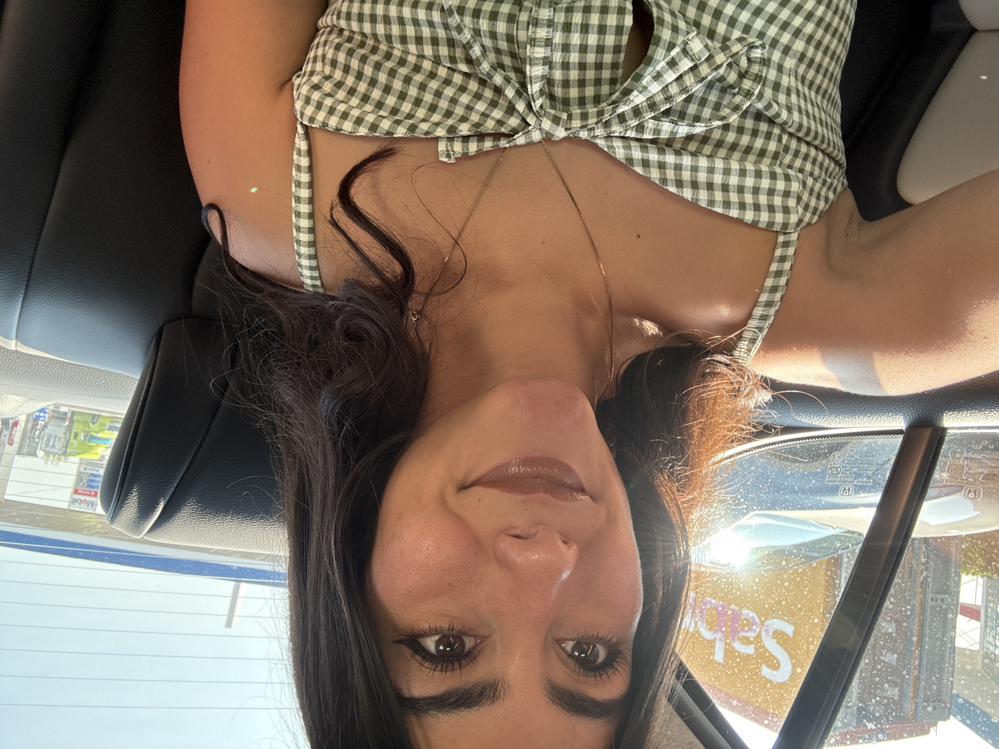 A medium shot captures a woman with dark hair and brown eyes wearing a green and white gingham top. She is in the driver's seat of a car. Outside the car window, a sign for "Sabritas" is visible, indicating the presence of this brand in the local environment, which appears to be in Santa Ana Xalmimilulco, Mexico.
