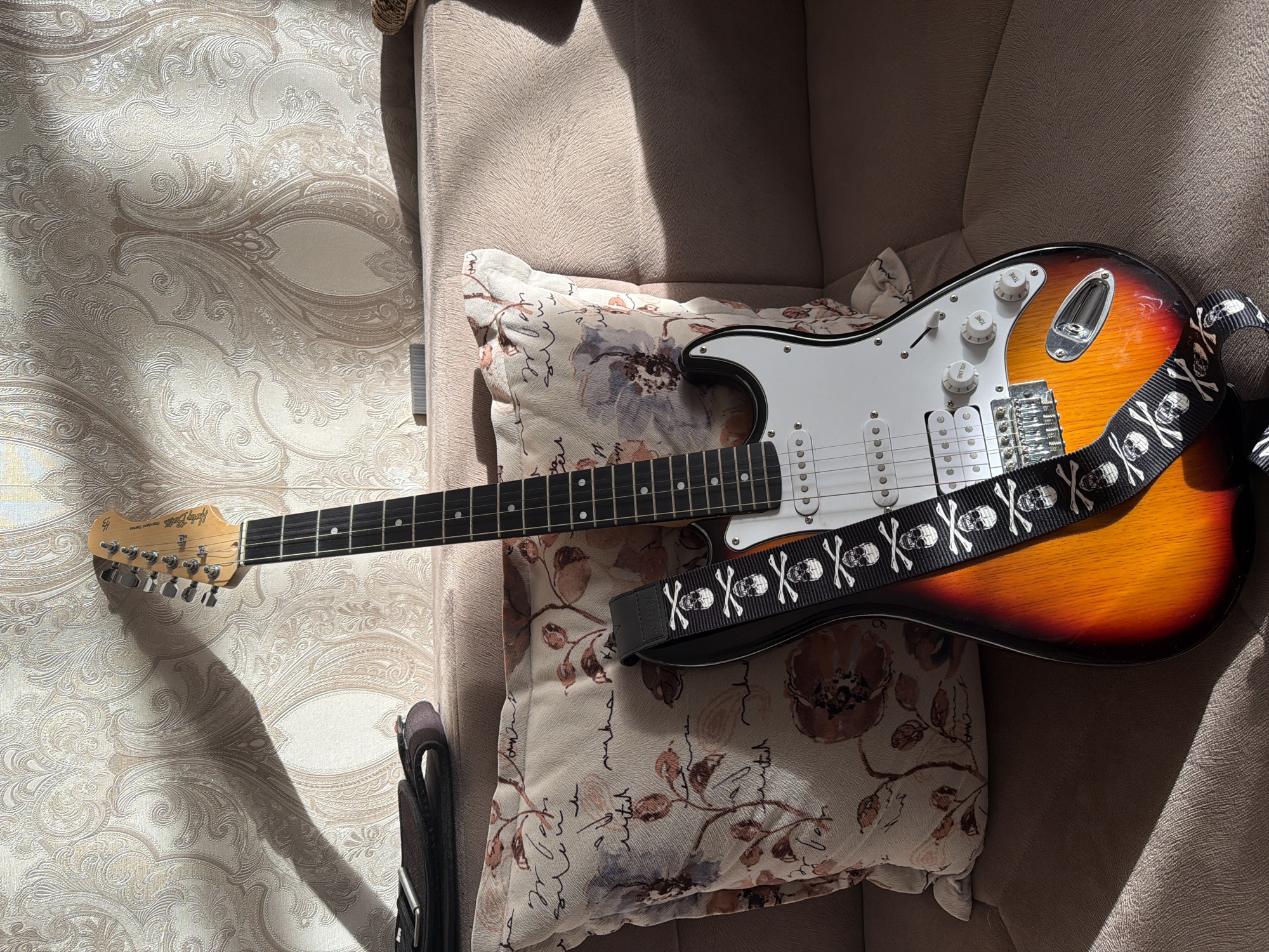The image shows an electric guitar resting on a couch and a pillow. The guitar is sunburst colored with a white pickguard and a black guitar strap adorned with skulls and crossbones. The guitar strap is clearly visible and takes up a significant portion of the frame. The couch appears to be made of a textured, light brown fabric. The pillow has a floral pattern with brown and blue flowers, and some faint script text. A wall with embossed wallpaper featuring a swirling, ornate design is visible to the left of the couch, and a shadow of the guitar's headstock is cast upon it. Sunlight streams in from the right side of the frame, creating distinct shadows and highlights on the couch and guitar, suggesting it's daytime. The overall scene is domestic and relaxed, with the focus on the guitar and its accessories. There are no people visible. The lighting and shadows suggest the sun is at an angle, possibly late morning or afternoon. The wallpaper and the style of the furniture provide some cues about the interior setting, but no specific location is identifiable beyond what is given in the prompt. There is text visible on the guitar headstock that appears to read "Squier Standard Series".