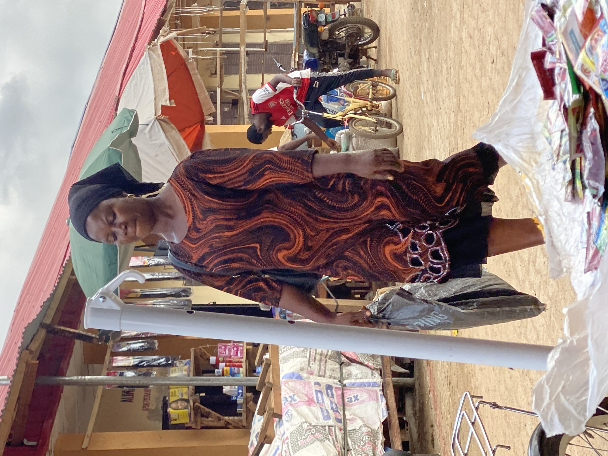 A woman wearing a brown and orange patterned dress and a black headscarf stands in what appears to be a market setting in Kafanchan B, Nigeria. She is positioned in front of a display of goods. In the background, a man wearing a red soccer jersey is seen repairing a bicycle. A motorcycle is parked nearby. The setting includes various market stalls with merchandise displayed, suggesting a bustling commercial environment. The overall image portrays a typical scene of daily life in a Nigerian market.