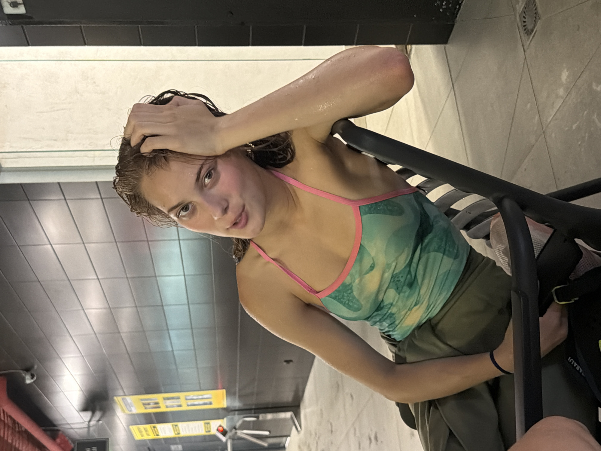 A young woman with wet hair is sitting on a black chair. She is wearing a green and yellow patterned tank top with pink trim and olive green pants. Her right arm is bent at the elbow, and her hand is touching her head. She is looking at the camera with her mouth slightly open. The background shows a tiled wall and some yellow signs with black text. The floor is tiled and appears to be wet in places. This scene is taking place in a public restroom in Kyiv, Ukraine, specifically within the Khreshchatyk metro station.