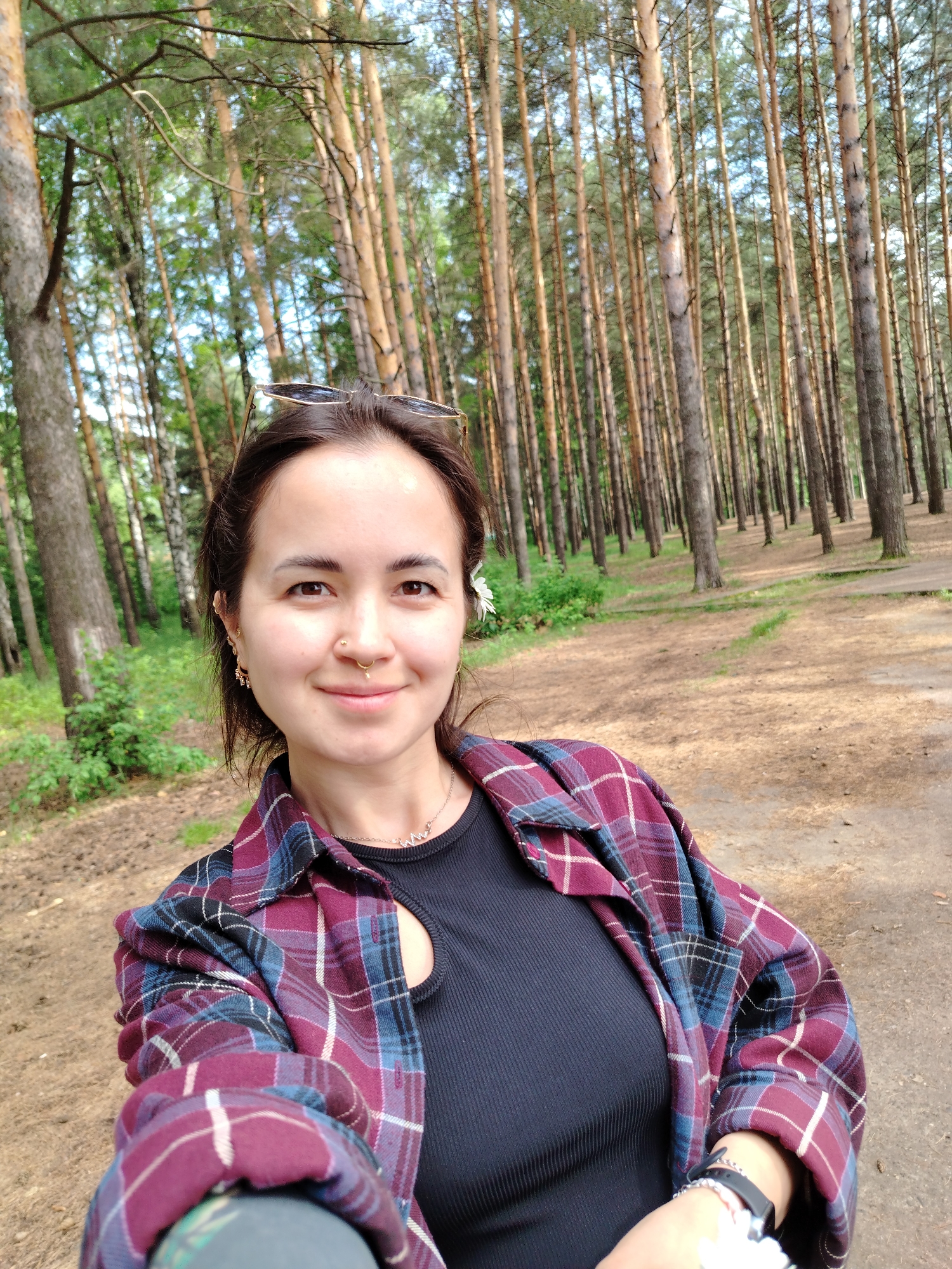 A smiling young woman takes a close-up selfie in a pine forest in Chaikovsky, Russia. She has dark hair, with sunglasses pushed onto her head and a small white flower tucked behind her right ear. Her expression is gentle and contented. Visible facial features include a septum piercing, a nostril piercing, and multiple earrings. She wears a black ribbed top and an open plaid shirt in shades of maroon, blue, black, and white. A silver necklace with a unique, angular charm is visible, as is a watch or bracelet on her left wrist.
The background shows numerous tall, slender pine trees with reddish-brown trunks and green foliage extending high into the sky, indicative of a dense forest. The ground is a light brown dirt path or clearing, suggesting a natural recreational area. The lighting is bright and natural, consistent with daytime. The image conveys a serene and pleasant outdoor atmosphere. No text is visible in the image.