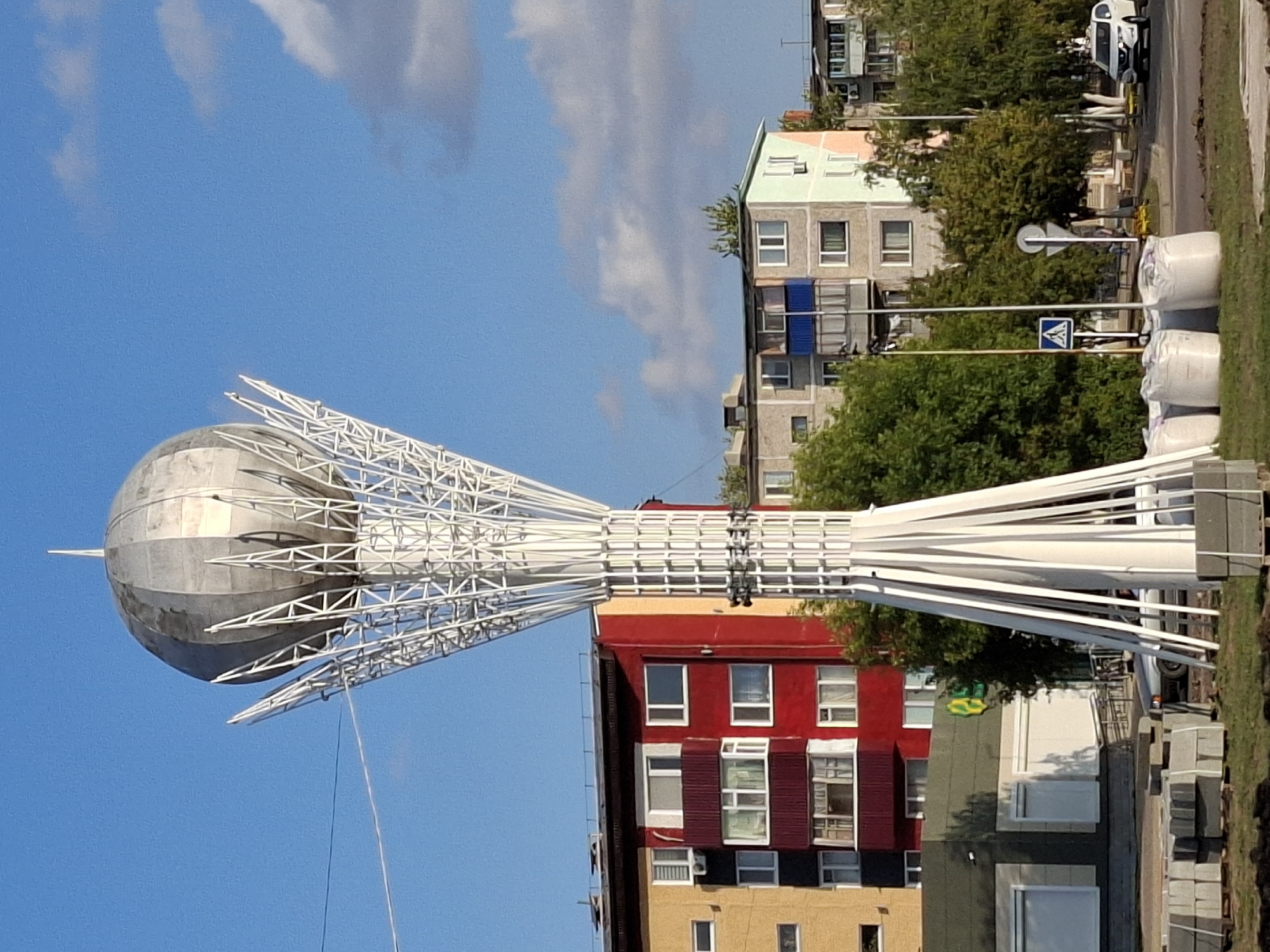 The image shows a tall, white, metallic sculpture resembling a blooming flower or a stylized tree with a spherical top. The sculpture is located in a city and is positioned in the foreground. Behind the sculpture, there are several apartment buildings. The closest building is painted in bright red and yellow. Further in the background, other apartment buildings of varying colors, including grey and pastel shades, are visible. Green trees are scattered throughout the scene, and a blue sky with some white clouds is overhead. A street with cars and a pedestrian crossing sign is also present. The base of the sculpture appears to be under construction, with piles of paving stones and large white sacks visible. The overall scene suggests an urban environment in Lisakovsk, Kazakhstan, likely a public space near residential buildings.