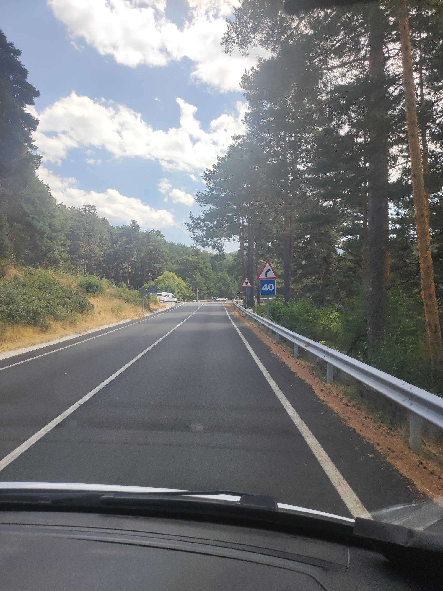 This is a first-person perspective, dashboard view of a paved road winding through a pine forest. The road has clear white lane markings, a white line on the right shoulder, and a metal guardrail. There are several road signs visible, including a speed limit sign indicating 40, and a curve warning sign. The day is sunny with scattered clouds. The surrounding environment consists of dense pine trees and undergrowth. A white van is parked on the side of the road in the distance. The scene appears to be on a road leading into or out of the city of Real Sitio de San Ildefonso, Spain.