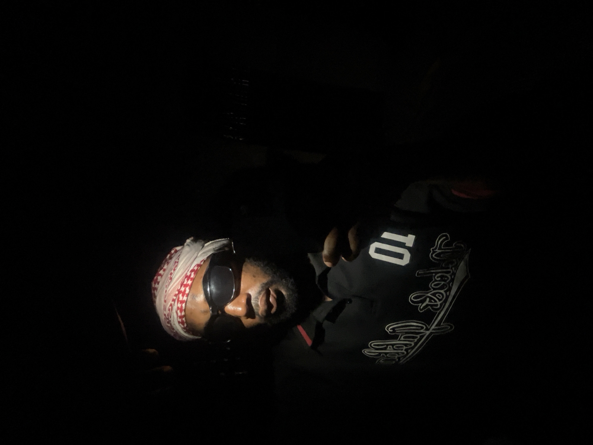 The image shows a man wearing sunglasses and a headscarf. He is wearing a black shirt with the number "10" on it and some other design on the side. It is difficult to ascertain the location of the image, though it has been designated as Lokoja, Nigeria. Due to the limited detail discernible, the time of day, environment, and setting cannot be accurately identified.