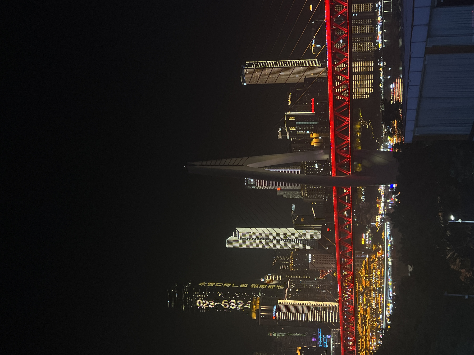 A nighttime cityscape of Jiangbei Cheng Street, China, features a prominent red suspension bridge illuminated with a lattice of lights, spanning across a body of water. On the left, a tall skyscraper displays a vertical illuminated advertisement with Chinese characters and the numbers "023-80321934". Adjacent to it, other buildings are lit up with varying patterns and colors. In the background, several more high-rise buildings are visible, some with horizontal or vertical lighting patterns. Boats with illuminated decks are seen on the water. The foreground is dominated by dark foliage and a concrete structure. The sky is dark and clear.